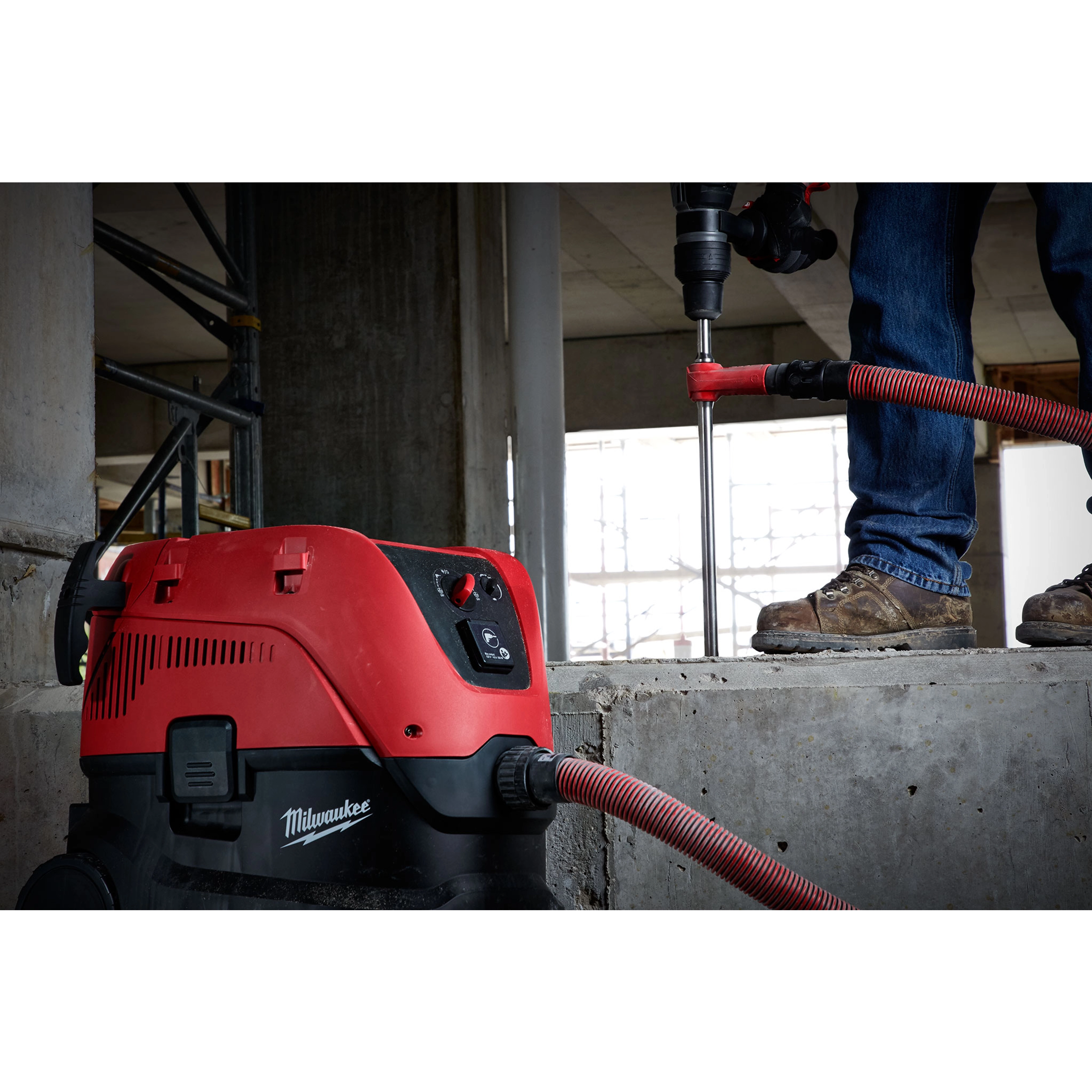A worker uses an SDS Vacuum Bit attached to a red and black vacuum cleaner to drill into a concrete surface at a construction site.