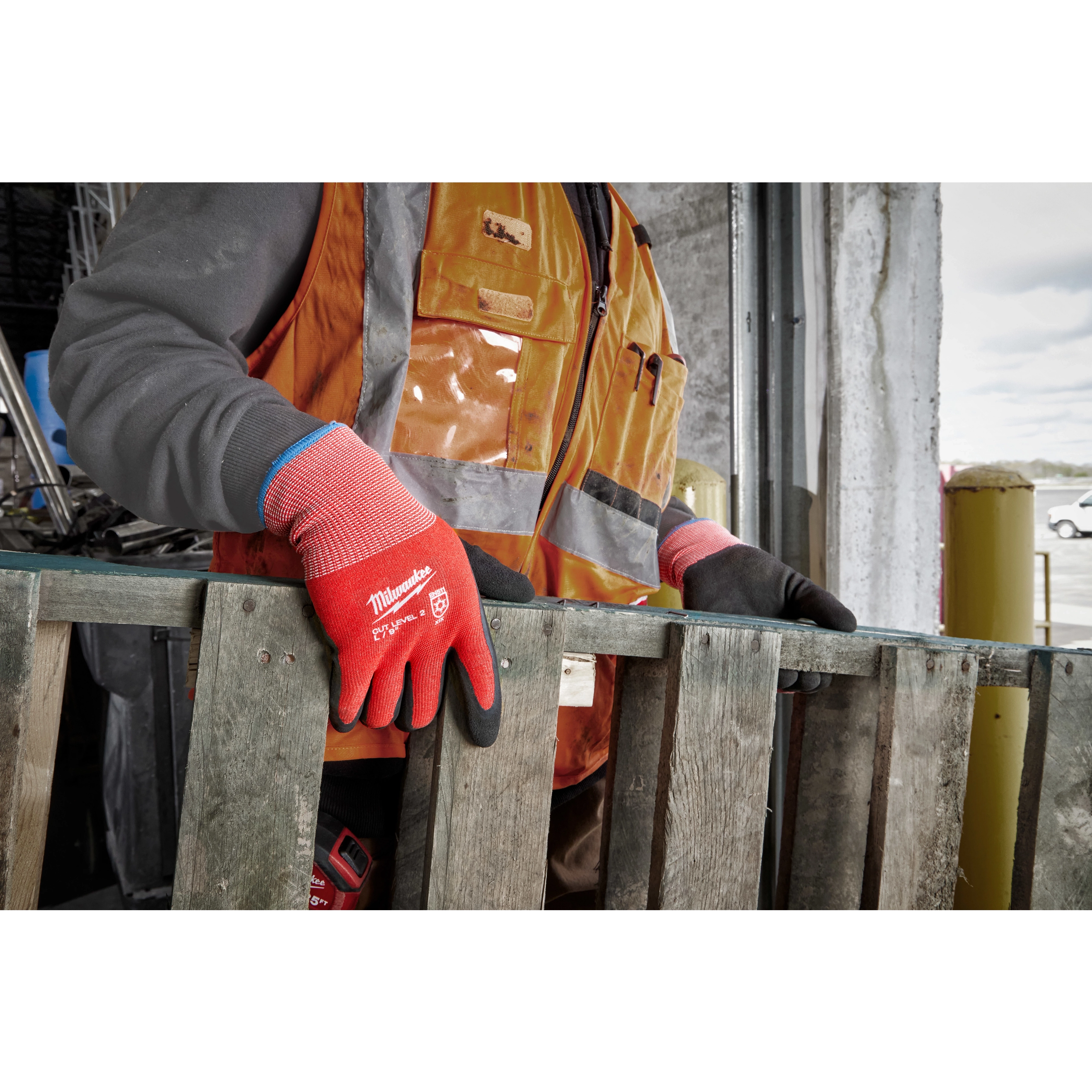 A worker wearing bright red Cut Level 2 Winter Dipped Gloves - L handles a wooden pallet. The worker is dressed in an orange safety vest and grey sleeves while standing in an industrial environment.