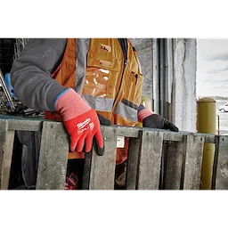 A worker wearing bright red Cut Level 2 Winter Dipped Gloves - L handles a wooden pallet. The worker is dressed in an orange safety vest and grey sleeves while standing in an industrial environment.