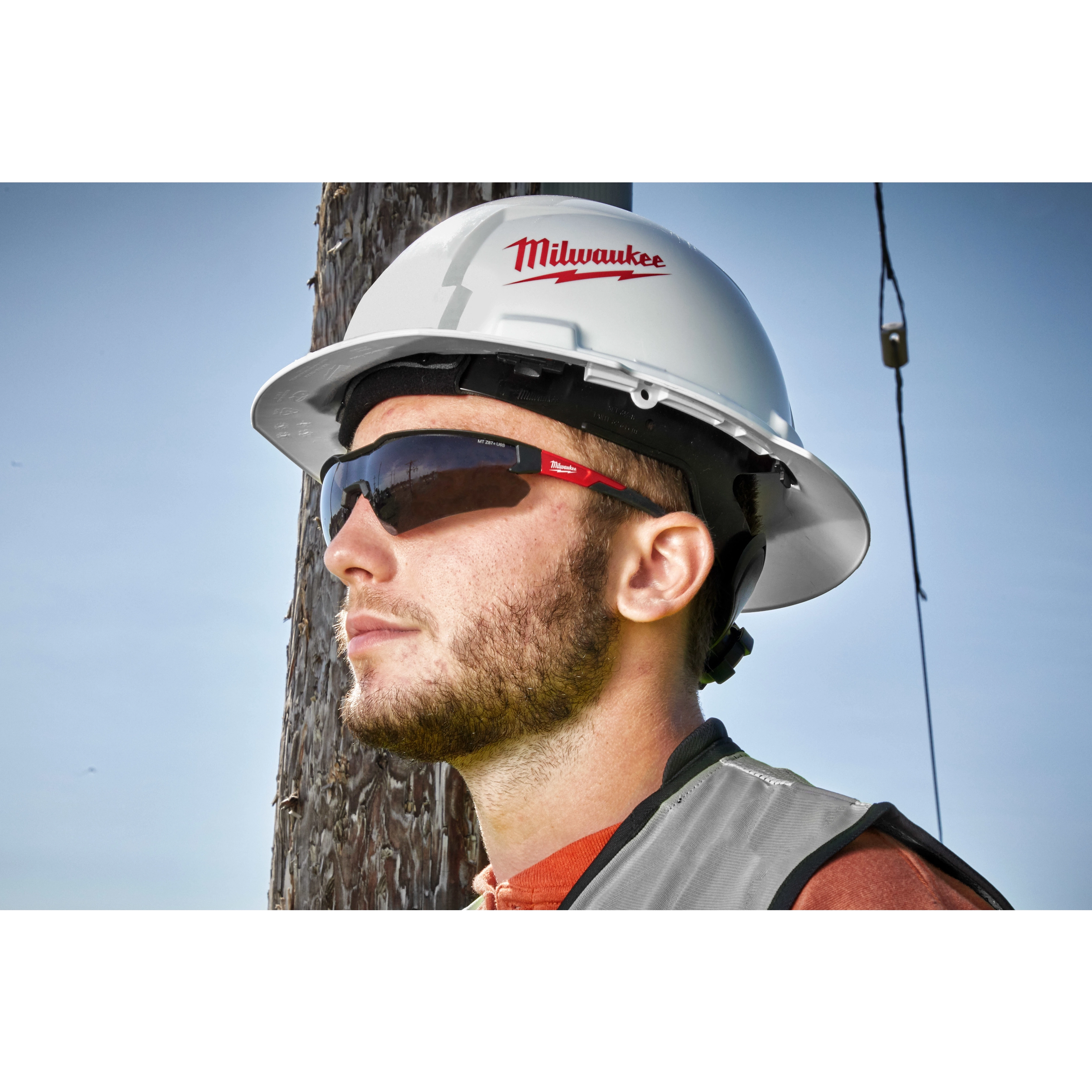 A person is wearing a white hard hat with the Milwaukee logo, a reflective vest, and Safety Glasses - Tinted Fog-Free Lenses, standing next to a utility pole.