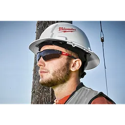 A person is wearing a white hard hat with the Milwaukee logo, a reflective vest, and Safety Glasses - Tinted Fog-Free Lenses, standing next to a utility pole.
