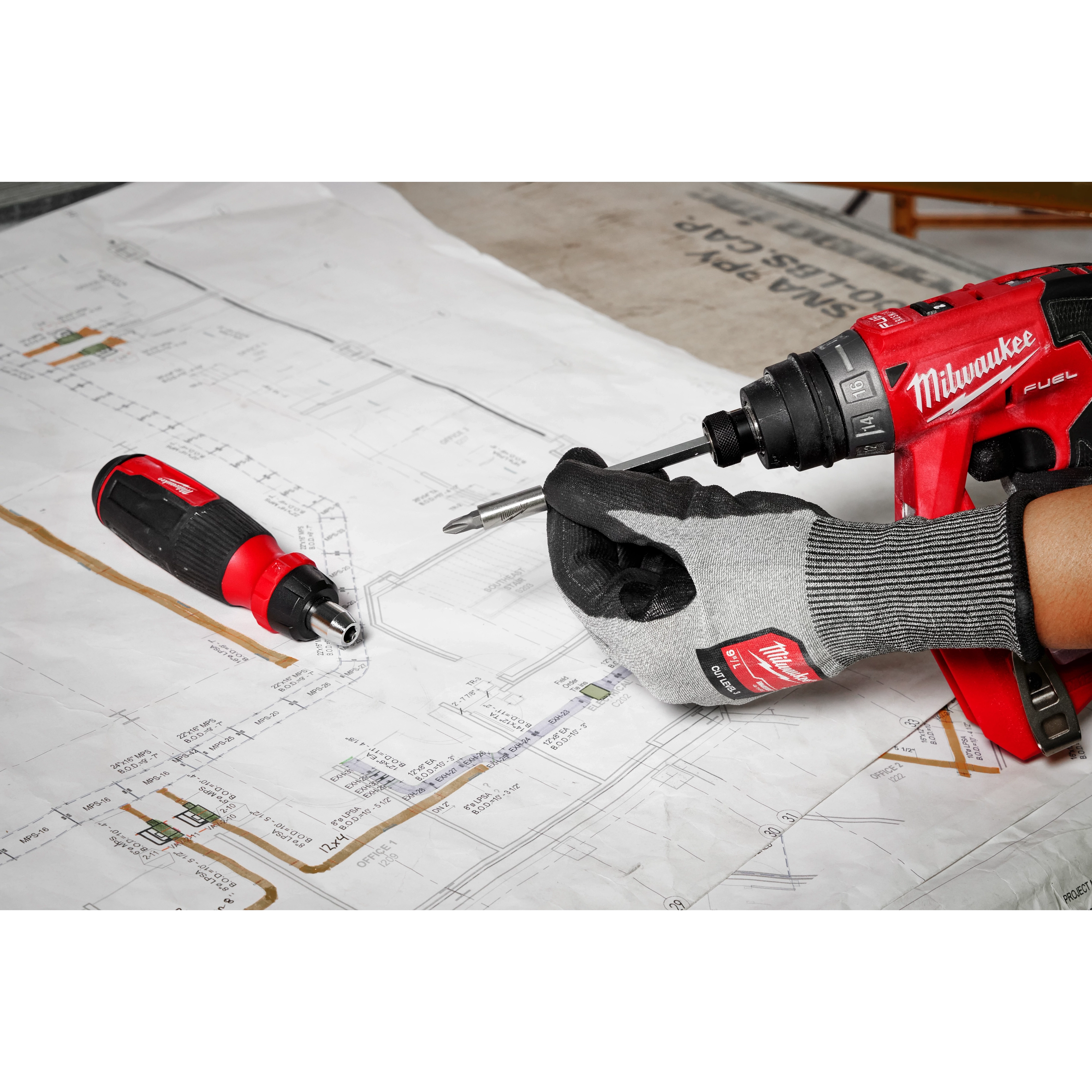 A gloved hand using a 14-in-1 Ratcheting Multi-Bit Screwdriver with a drill over architectural blueprints, with another screwdriver lying nearby.