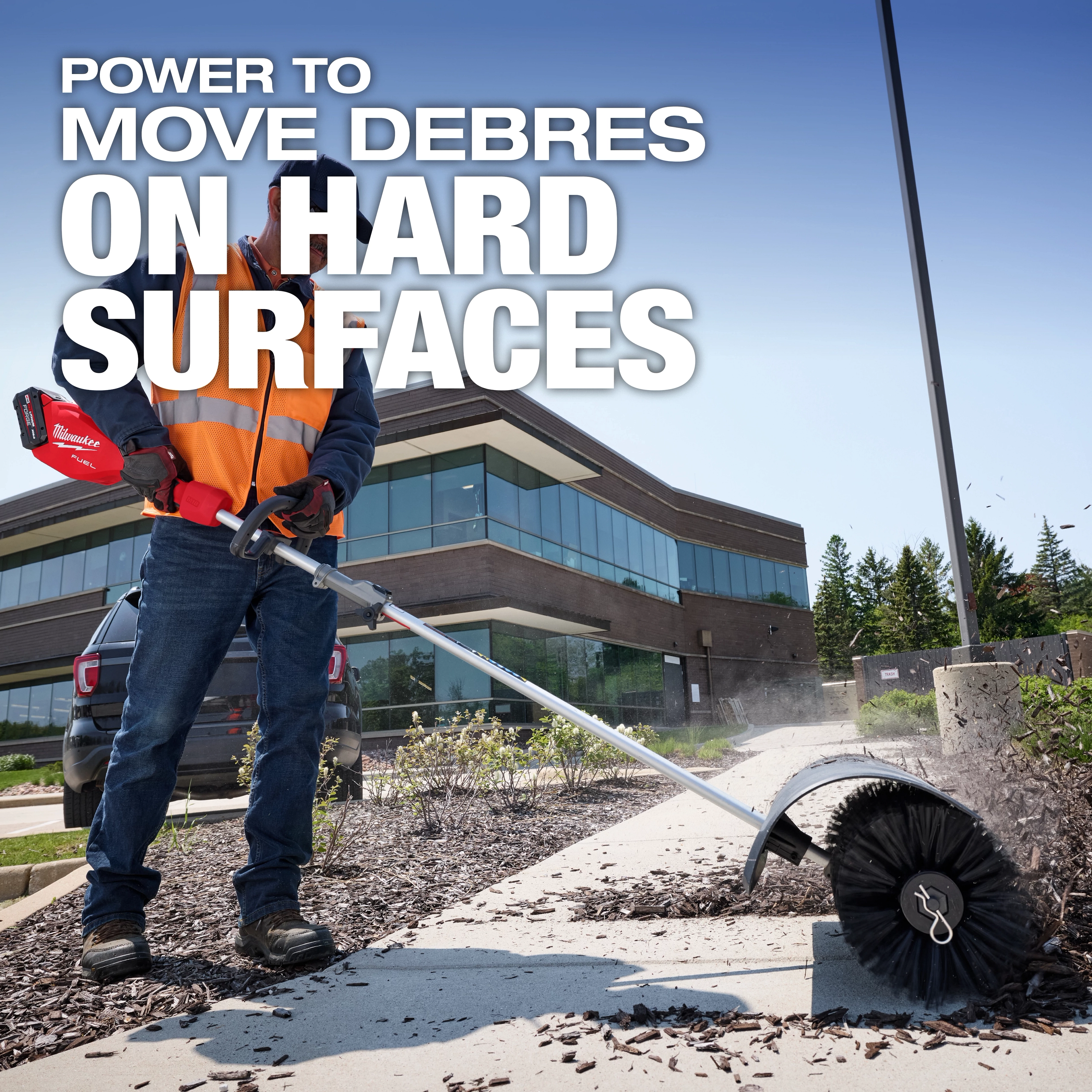 Worker using M18 FUEL QUIK-LOK Bristle Brush Attachment to clear debris from a sidewalk near a building.