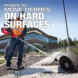 Worker using M18 FUEL QUIK-LOK Bristle Brush Attachment to clear debris from a sidewalk near a building.