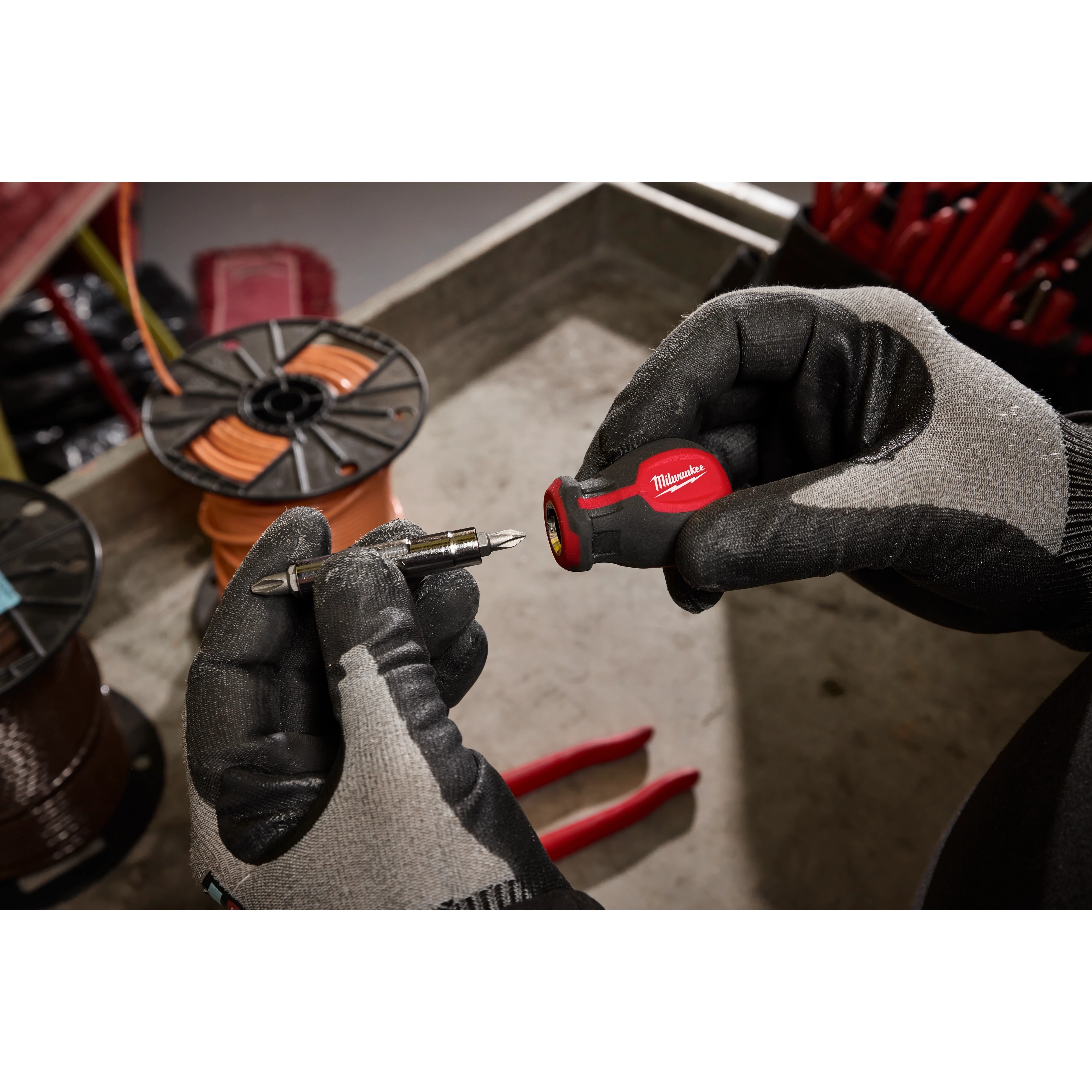 A person wearing gloves is holding a 6-IN-1 Stubby Multi-Bit Screwdriver in one hand and a detachable bit in the other. The background includes tools and wire spools.