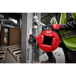 Worker using a red 1/4" 100' Steel Fish Tape while wearing safety gear and gloves in an industrial setting, holding the tape near pipes.