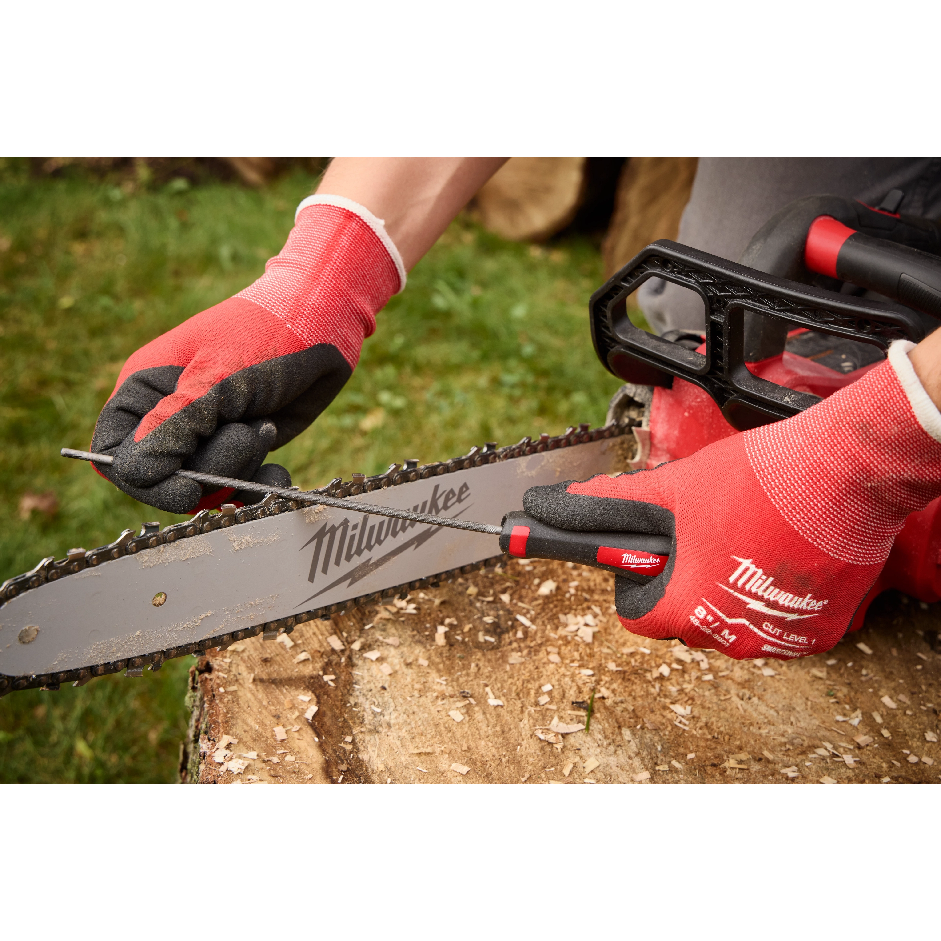 Person using a file to sharpen the chain of a Milwaukee chainsaw. They are wearing red and black Milwaukee gloves and the chainsaw is resting on a tree stump with green grass in the background.
