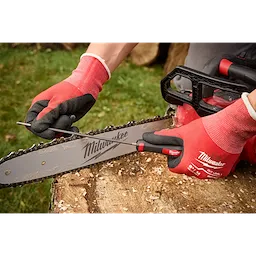 Person using a file to sharpen the chain of a Milwaukee chainsaw. They are wearing red and black Milwaukee gloves and the chainsaw is resting on a tree stump with green grass in the background.