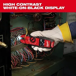 A gloved hand holds the Milwaukee multimeter in front of an electrical panel, testing wires. The multimeter features high contrast white-on-black display for easy reading. The surrounding area has colorful exposed wires and components, illustrating the tool's application in an electrical maintenance setting.