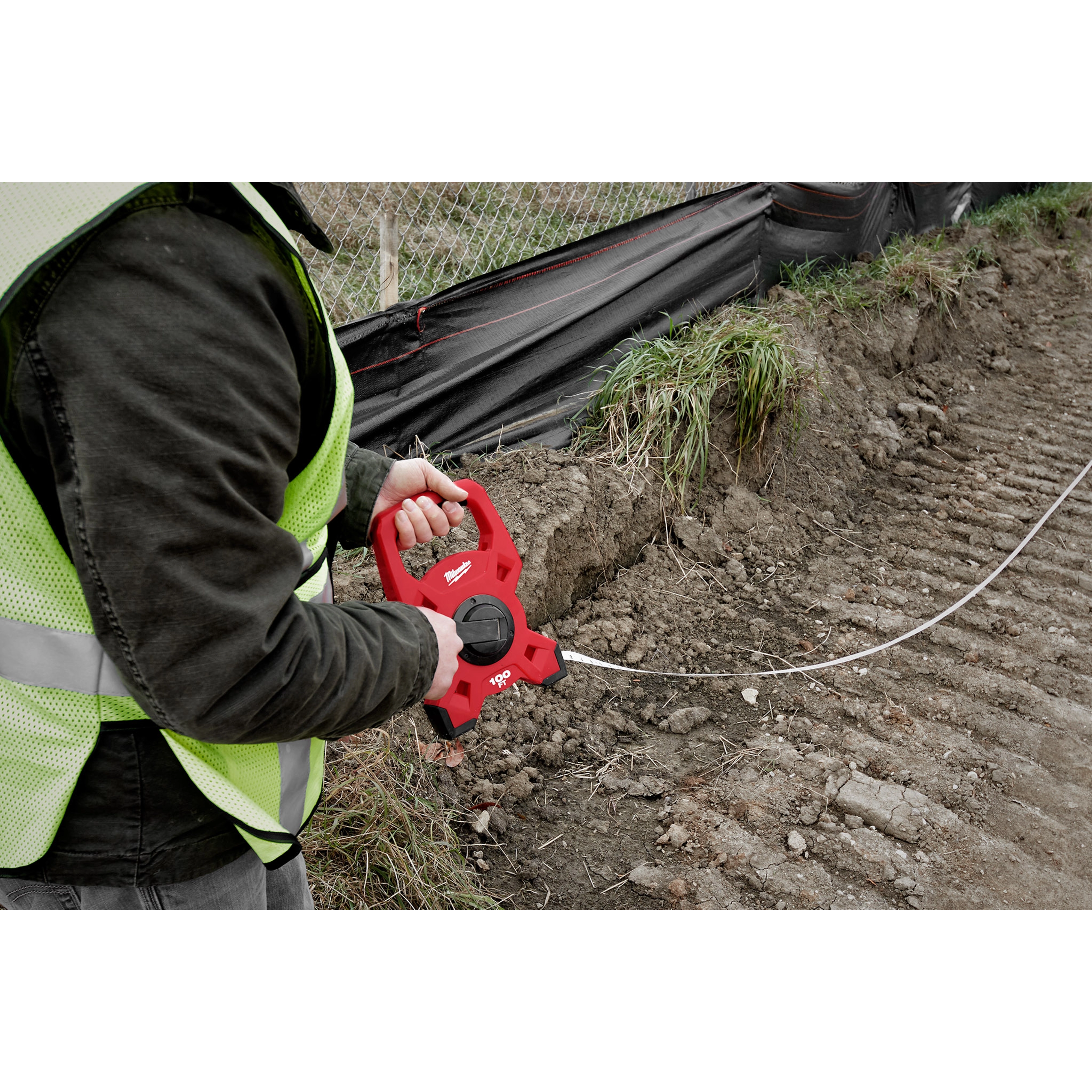 A person holds a Milwaukee 100 Foot Fiberglass Long Tape at a construction site, preparing to measure the ground.