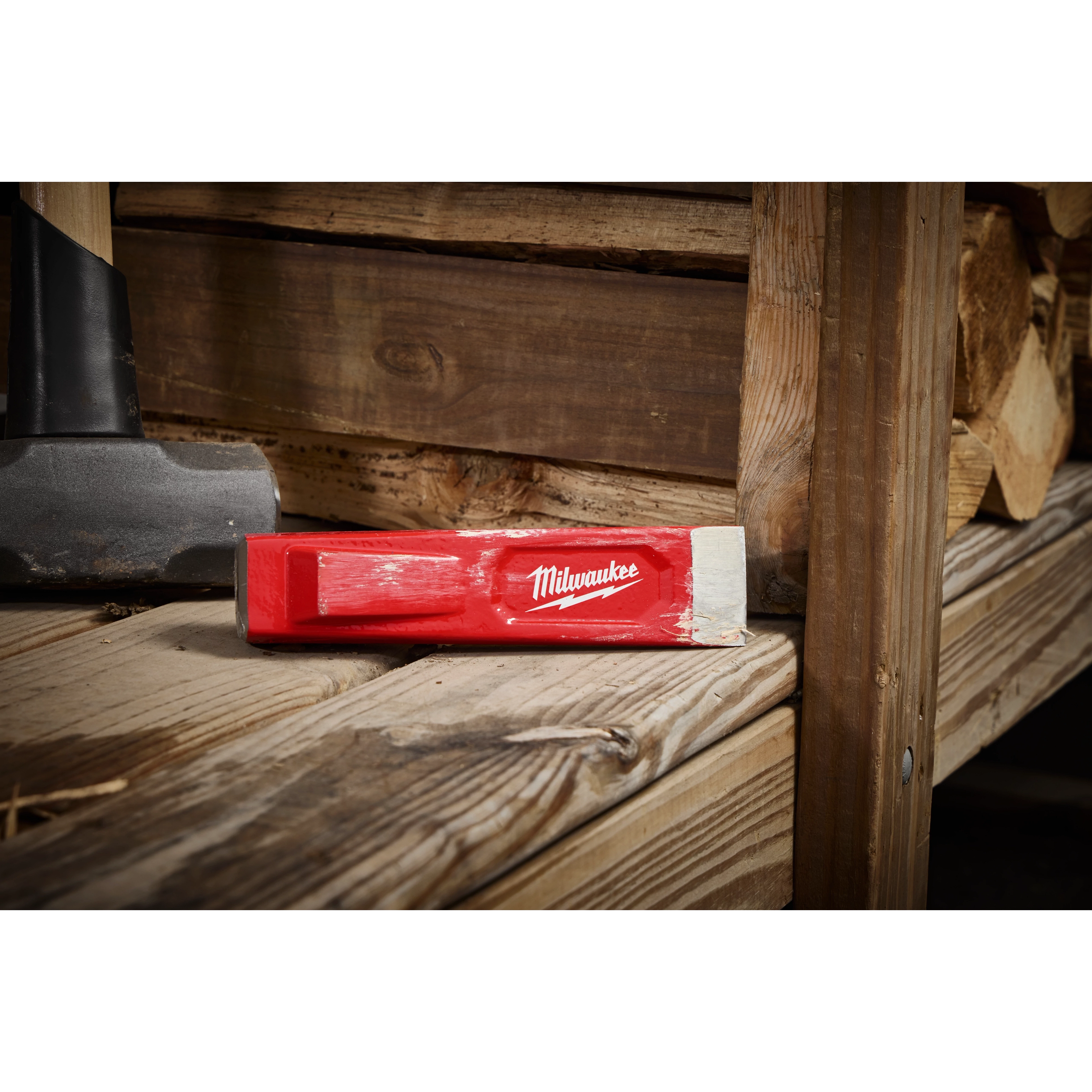 The 5lb Splitting Wedge with a red coating and the Milwaukee logo is placed on a wooden surface next to a weathered sledgehammer in a rustic setting.