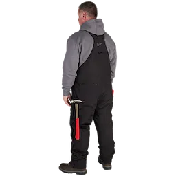 Man wearing FREEFLEX Insulated Bib Overalls in black, grey hoodie, while holding a hammer with a red handle, standing outdoors.
