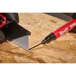 The INKZALL™ 2IN1 Deep Reach Ultra Fine Point Black Jobsite Marker is used to mark a precise line on a piece of wood next to a utility knife blade. The marker has a red body, and both hands wear black gloves.