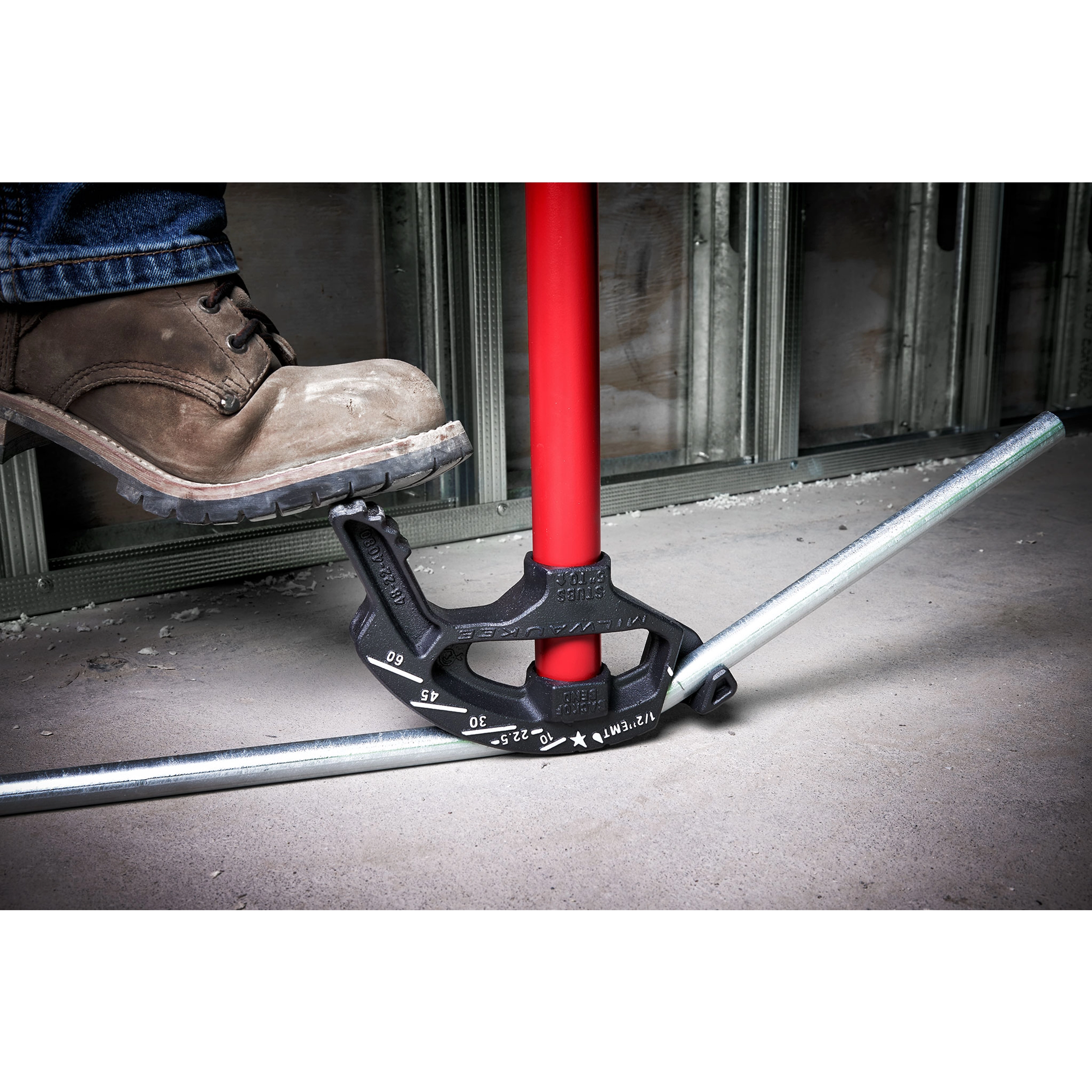 A worker bends a metal conduit using a 1/2 EMT Iron Conduit Bender with a red handle. The foot pressing down aids the bending process.