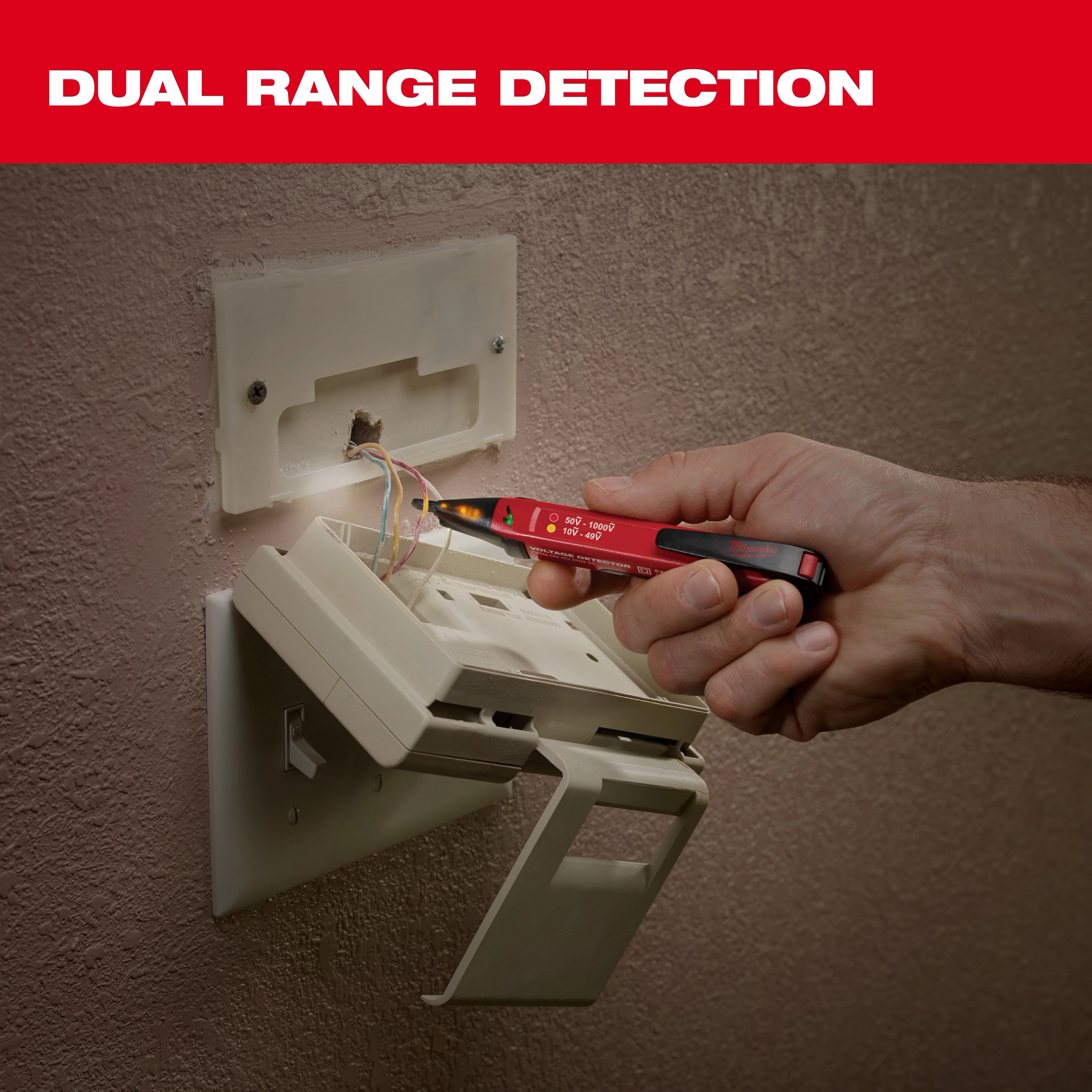 A hand uses the Milwaukee Dual Range Detection tool to test wires in a wall outlet box. The red pen-shaped tool has indicators for detecting voltage in the wires. A caption above reads "DUAL RANGE DETECTION" in white text on a red background.