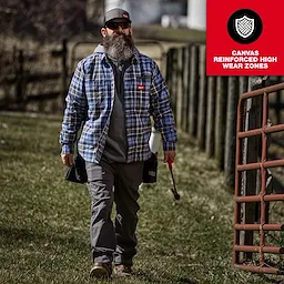 A person wearing a GRIDIRON Flannel Shirt walks outdoors carrying a tool and a bag. The shirt features canvas reinforced high wear zones.