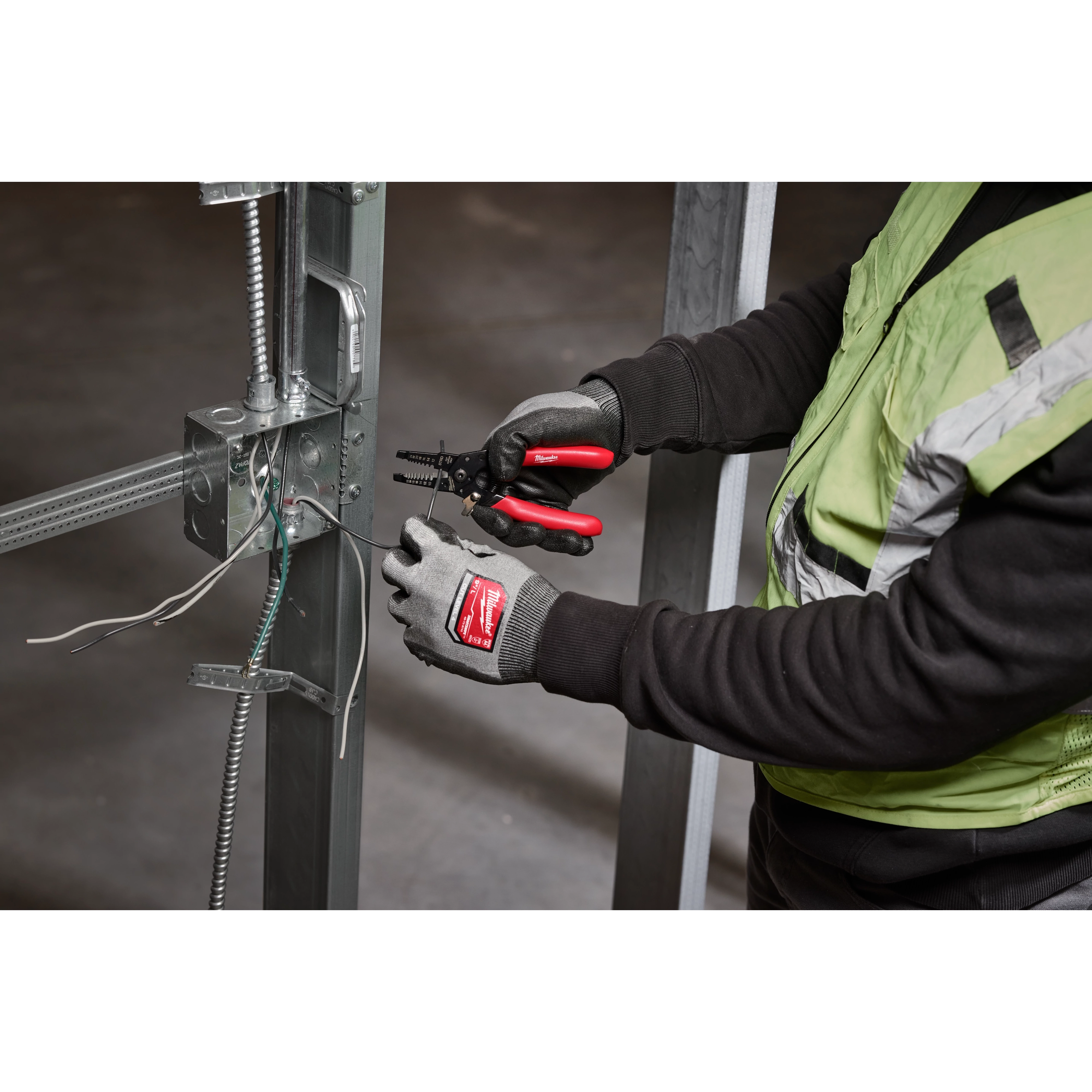 A person wearing gloves and a safety vest uses a 10-24 AWG Compact Dipped Grip Wire Stripper & Cutter to work on electrical wires attached to a metal structure inside an unfinished building.