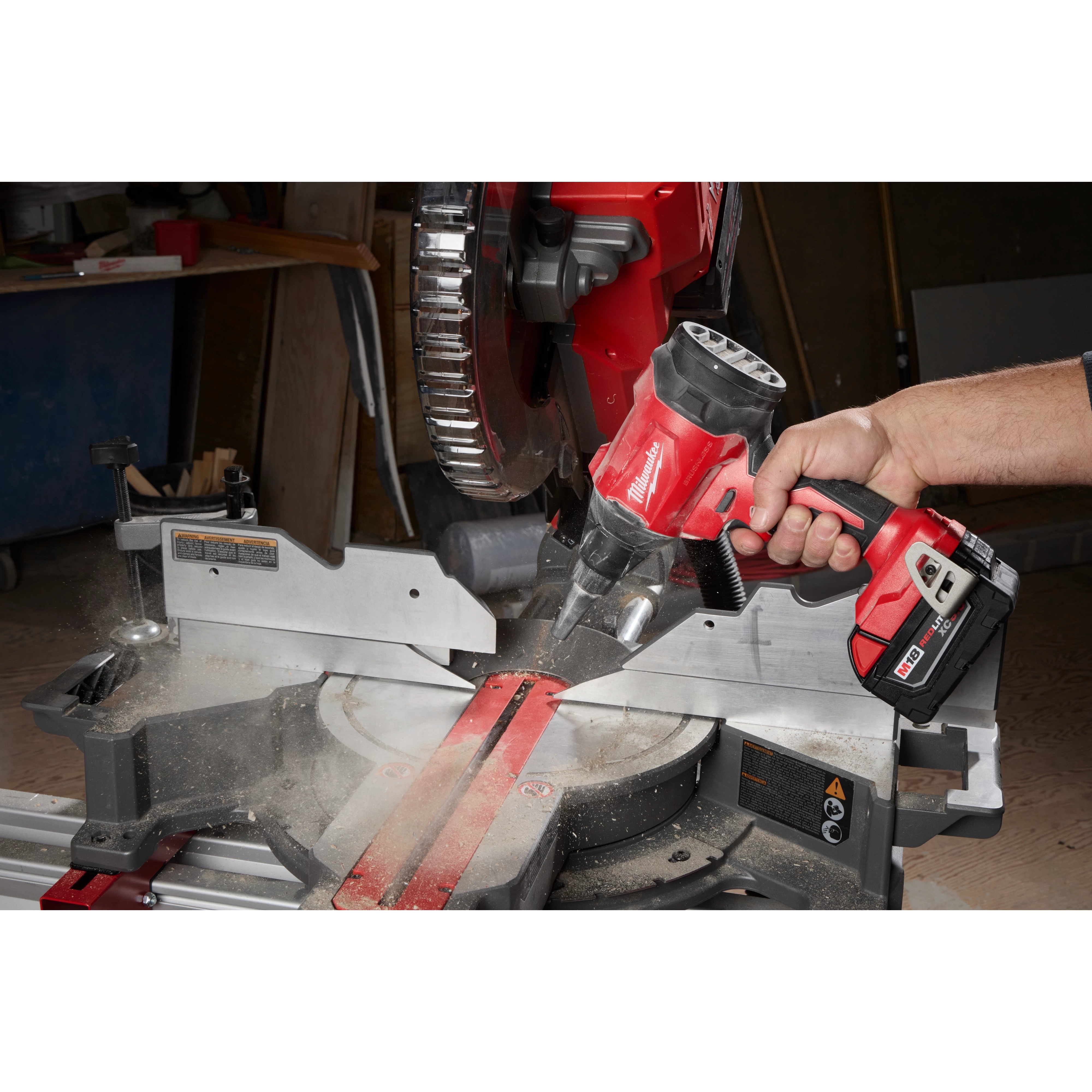 A person's hand uses the M18™ Brushless Precision Blower to remove sawdust from a miter saw in a workshop. The blower is red and black with a compact design, enhancing cleaning efficiency around tools.