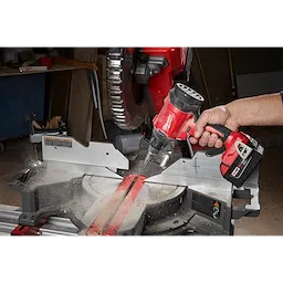 A person's hand uses the M18™ Brushless Precision Blower to remove sawdust from a miter saw in a workshop. The blower is red and black with a compact design, enhancing cleaning efficiency around tools.