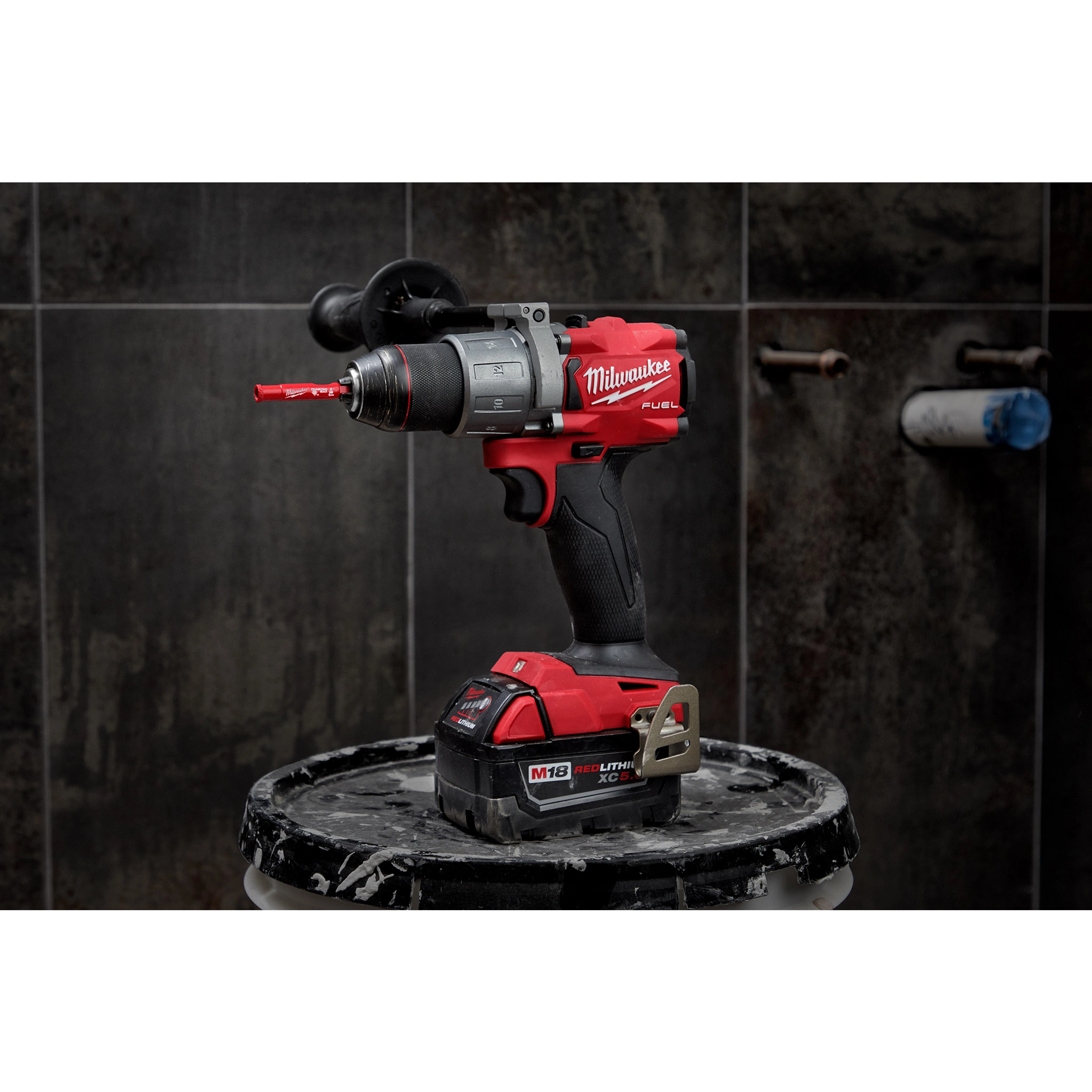 Power drill with a 1/4" Diamond Grit Hole Saw attachment, placed on a paint-splattered stool against a dark tiled wall background.