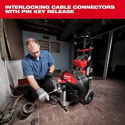A man wearing gloves operates a Milwaukee cable connector machine in a basement. The machine features interlocking cable connectors with a pin key release, designed for heavy-duty cable work and ease of use. It has red and black components, and is equipped with wheels for mobility.