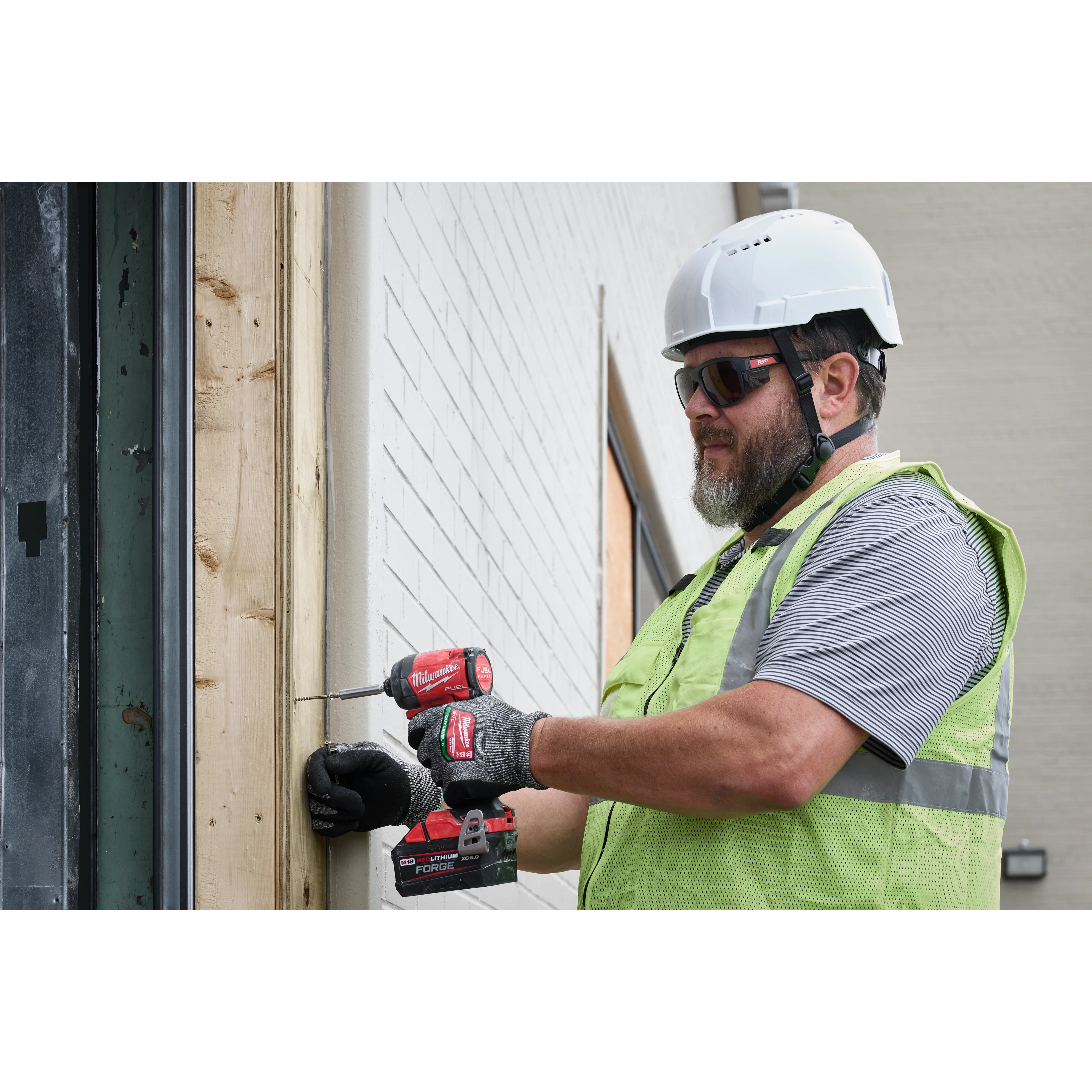 A construction worker wearing Full Frame Safety Glasses with Removable Side Shields – Silver Mirrored Anti-Scratch Lenses is using a red and black power drill to secure a screw into a wooden beam. The worker also wears a white hard hat, black gloves, and a green safety vest.