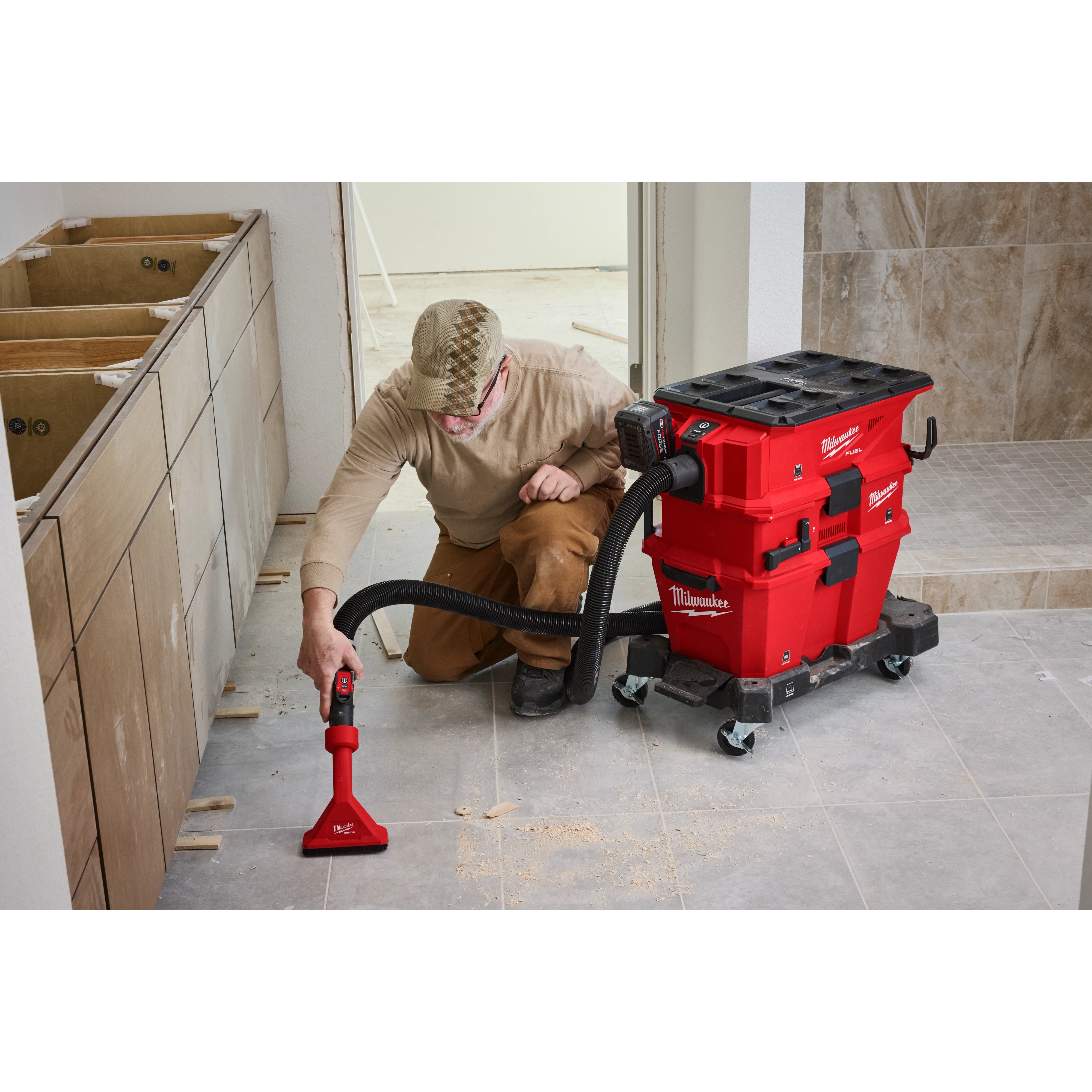 A person wearing a cap and protective eyewear uses a NEXUS™ Dedicated Filter Cleaner to clean up debris on a tiled floor in an under-construction room. The cleaner is red and black, with a flexible hose and attachments attached to it.