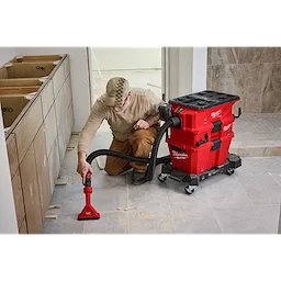 A person wearing a cap and protective eyewear uses a NEXUS™ Dedicated Filter Cleaner to clean up debris on a tiled floor in an under-construction room. The cleaner is red and black, with a flexible hose and attachments attached to it.