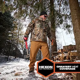 A person wearing the M12 Heated QUIETSHELL Jacket stands outdoors in a snowy setting, holding a tool with a pile of chopped wood nearby.