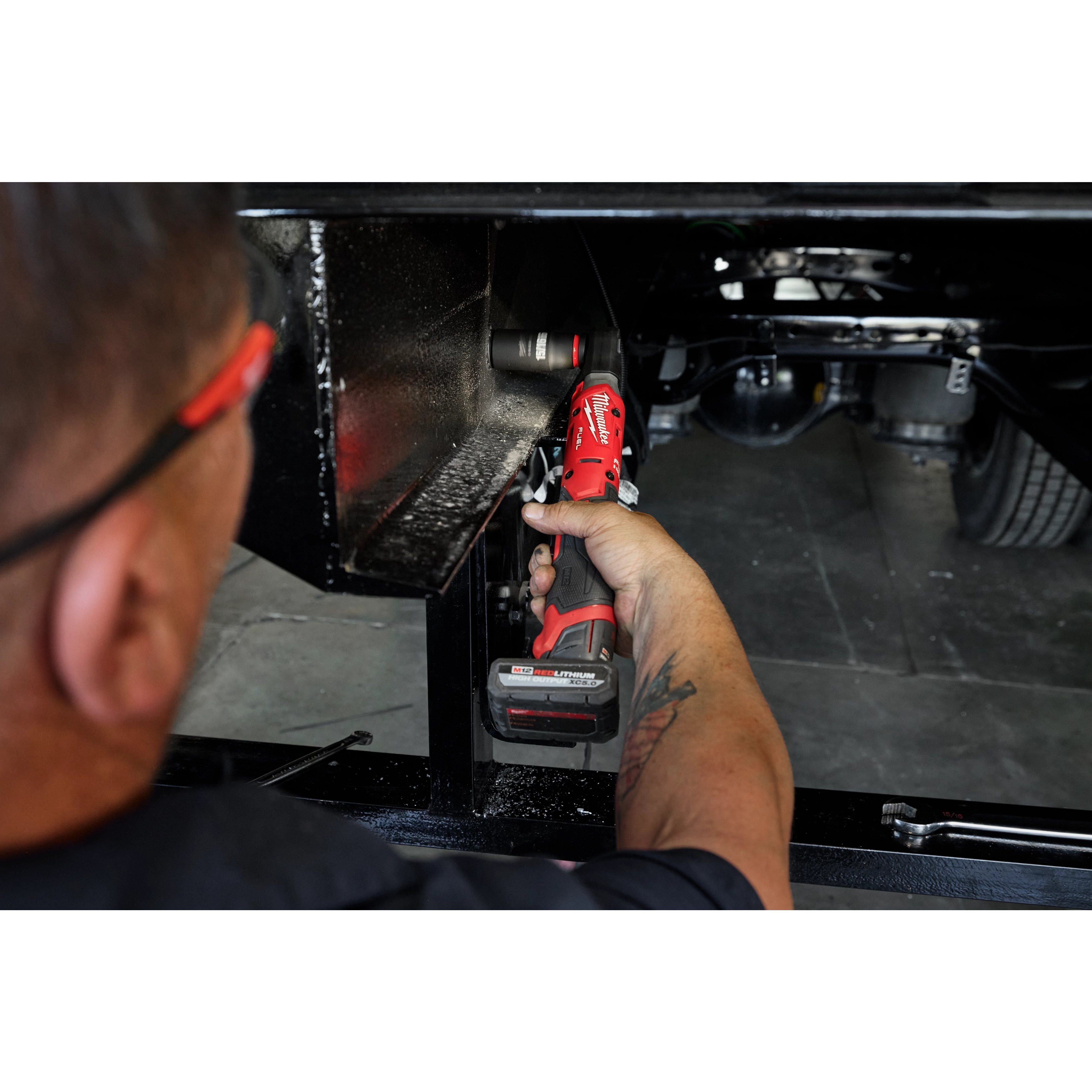 A person uses the M12 FUEL™ 1/2" Ratchet Kit to tighten a bolt in a difficult-to-reach area under a vehicle. The red and black ratchet features a battery pack and ergonomic handle.