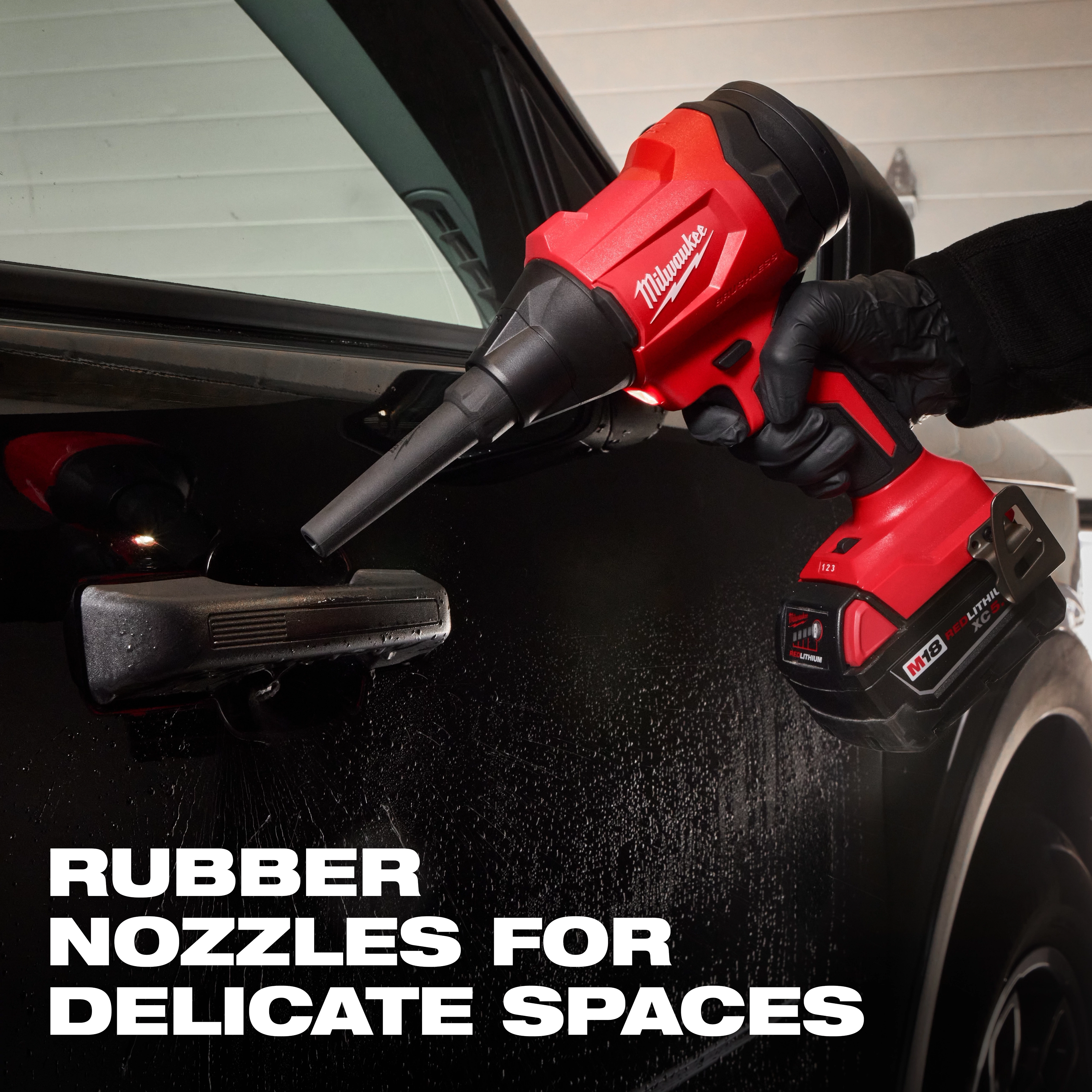 A person wearing gloves is using the red and black M18™ Brushless Precision Blower by Milwaukee to clean delicate spaces around a black car door's handle. Text on the image reads, "RUBBER NOZZLES FOR DELICATE SPACES."