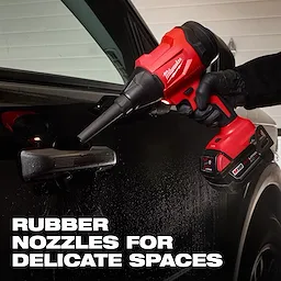 A person wearing gloves is using the red and black M18™ Brushless Precision Blower by Milwaukee to clean delicate spaces around a black car door's handle. Text on the image reads, "RUBBER NOZZLES FOR DELICATE SPACES."