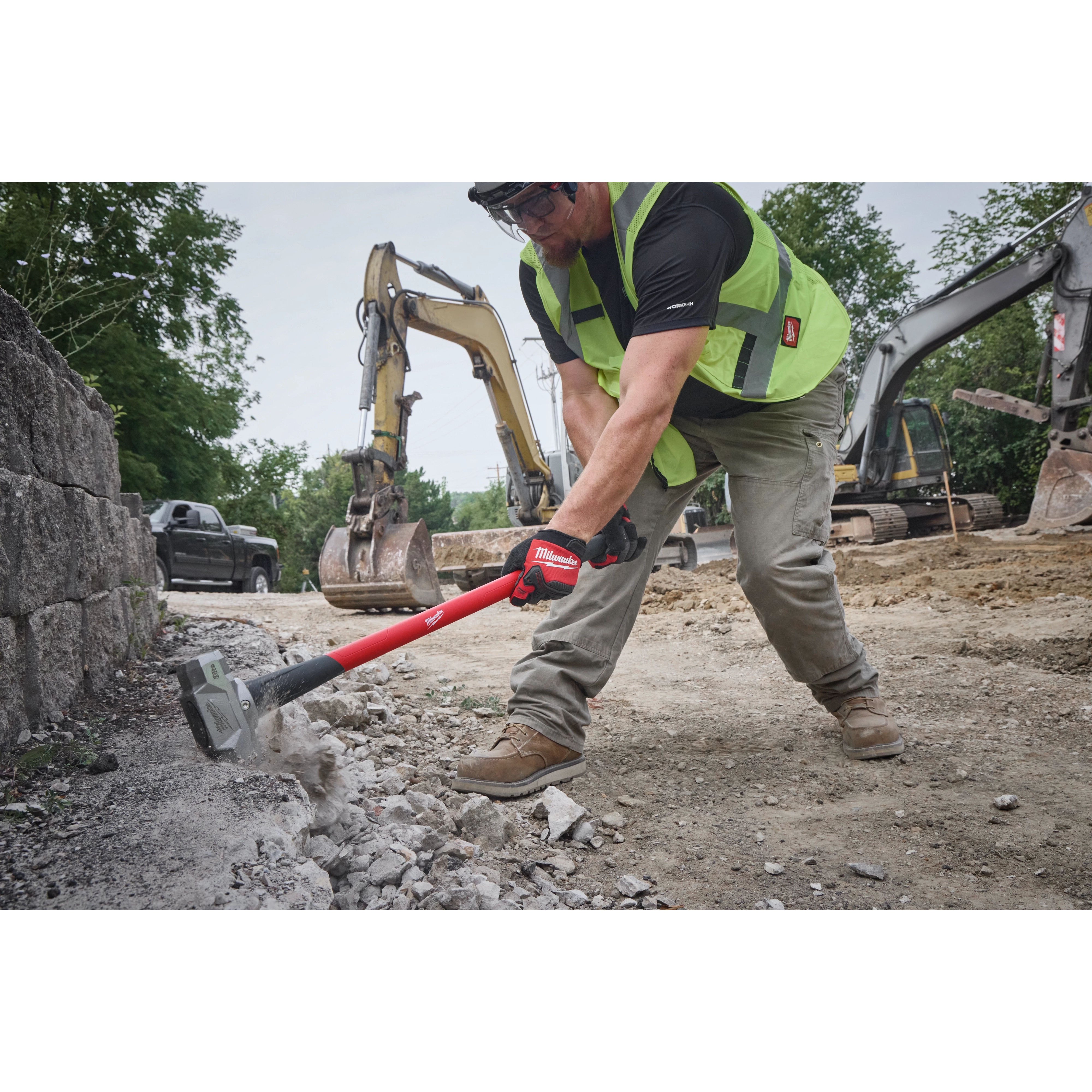 A construction worker is using an 8-lb Sledge Hammer (36” Handle) to break up concrete on a job site. The hammer has a red and black handle, and excavators and trucks are in the background on the dirt. The worker wears gloves, a safety vest, and work boots.