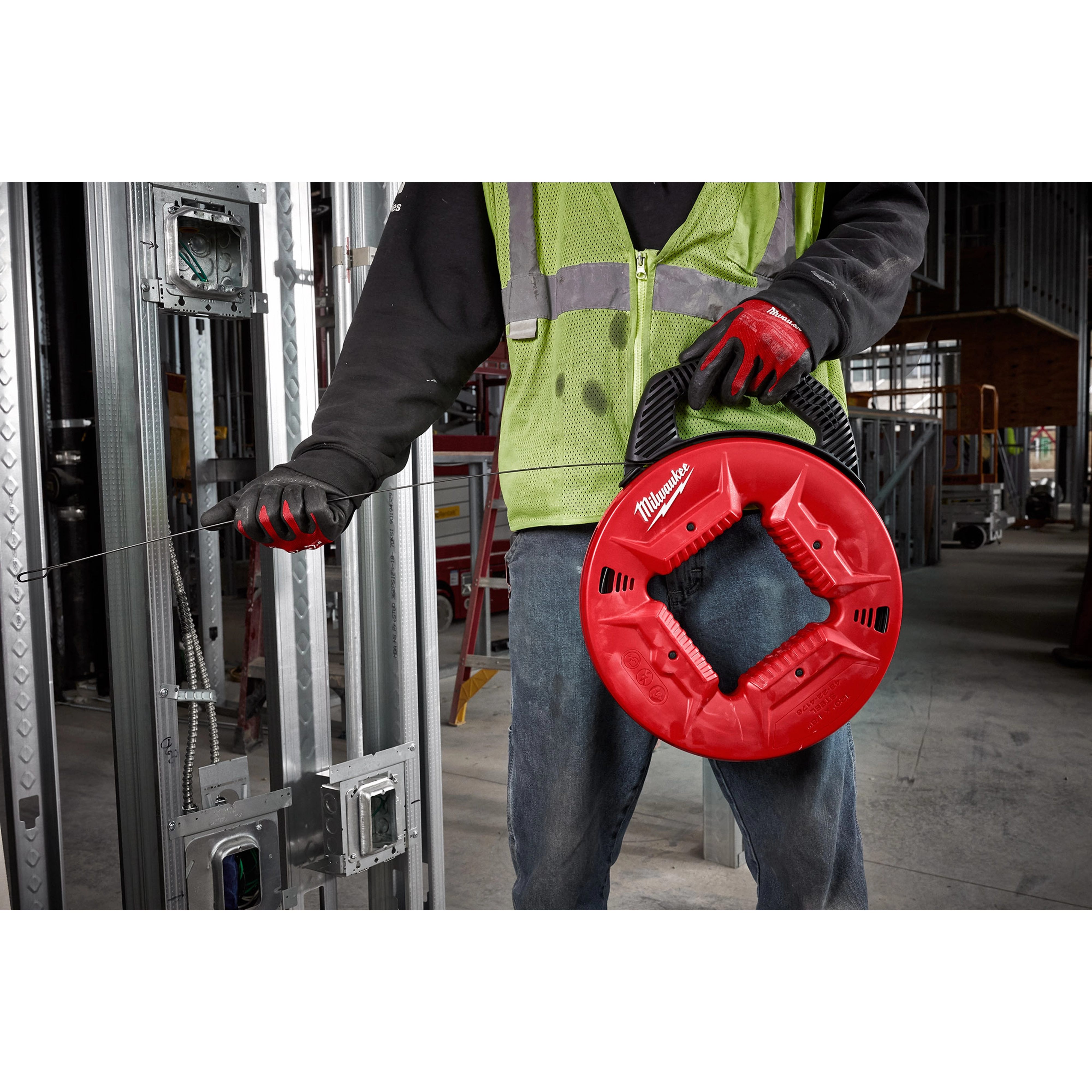 Person using 1/8" 120' Steel Fish Tape in a red reel, wearing a green safety vest and red gloves, at a construction site.