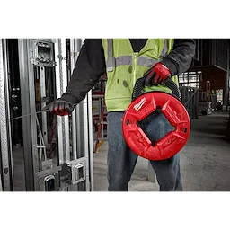 Person using 1/8" 120' Steel Fish Tape in a red reel, wearing a green safety vest and red gloves, at a construction site.