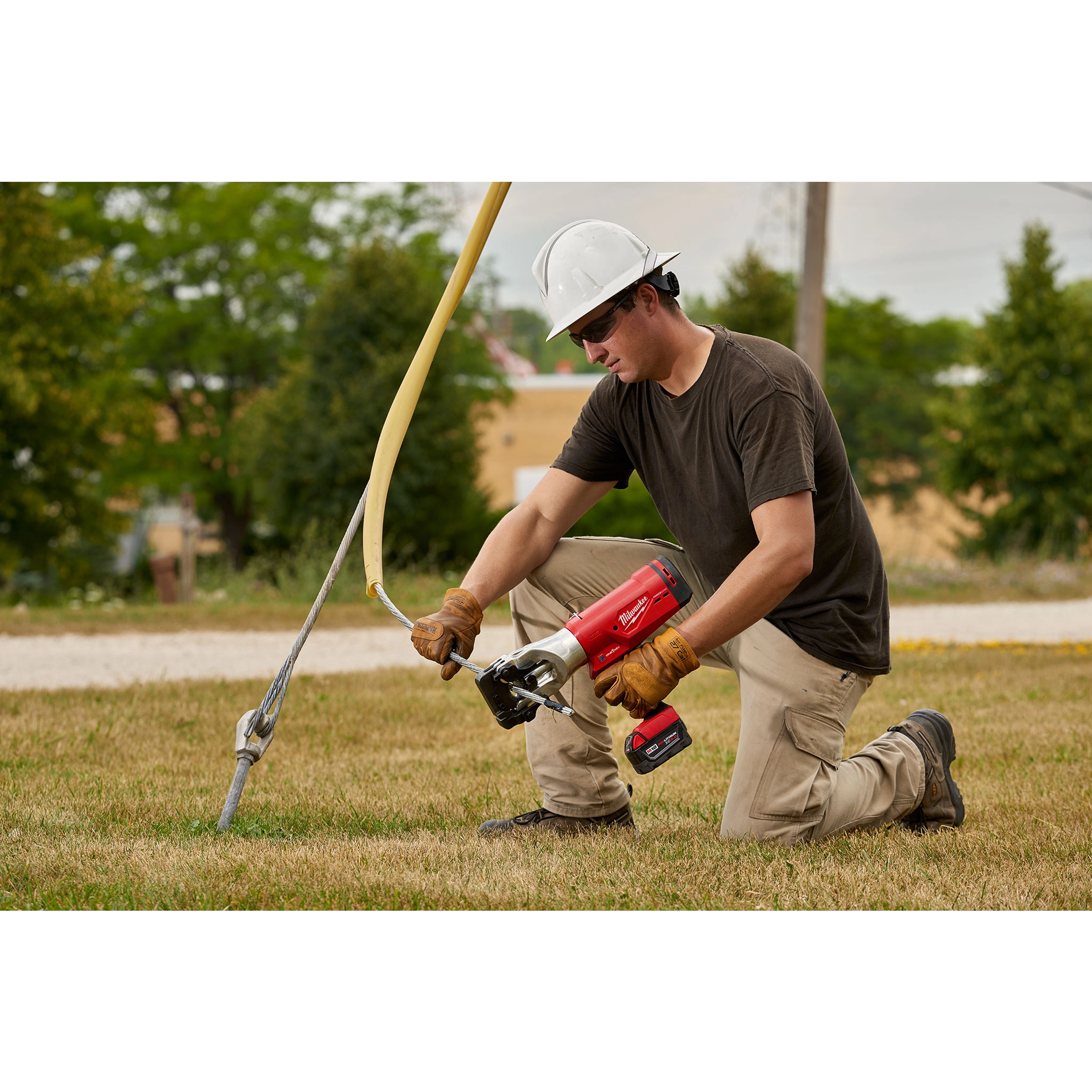 A worker using the FORCELOGIC™ M18™ 1590 ACSR Cutter Kit to cut a wire secured to a pole. The worker is wearing a white hard hat, brown short-sleeve shirt, and beige pants, while kneeling on grass.