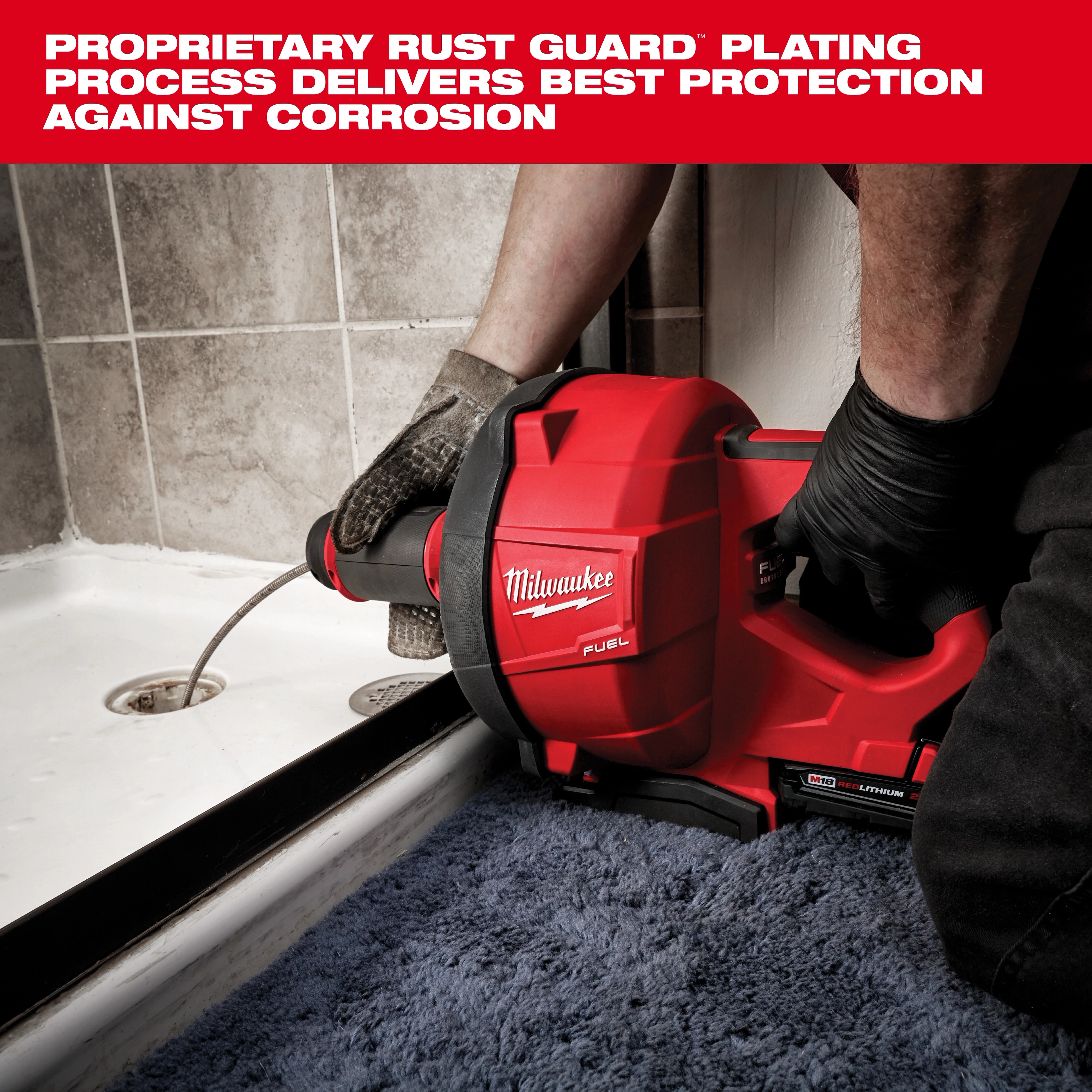 A Milwaukee Fuel drain cleaner is in use, with a worker's gloved hands guiding the cable into a shower drain. The red tool is prominently labeled with the brand name and is designed to prevent corrosion with its rust guard plating. The area surrounding the drain is tiled and has a blue carpet.