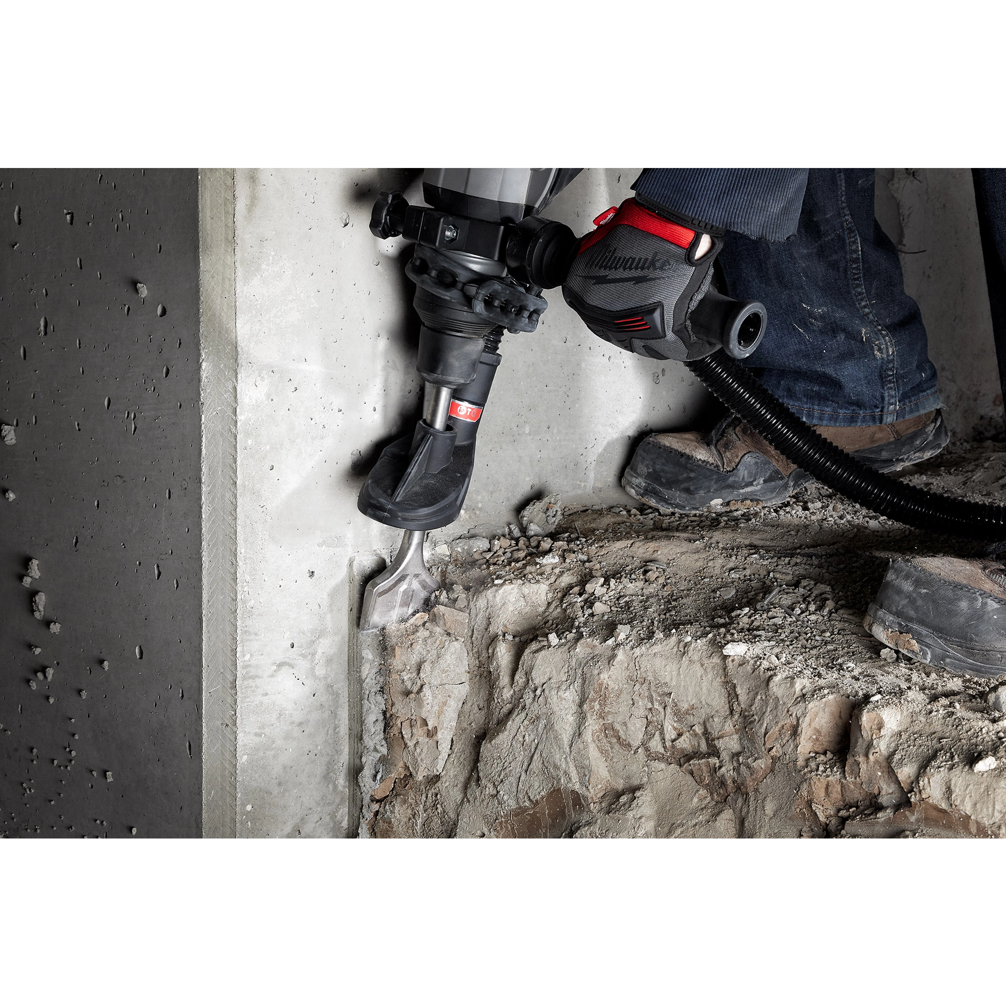 Person using the SDS-MAX SLEDGE 3" SCALING CHISEL with a hammer drill to remove concrete from a wall. Debris and dust are visible.