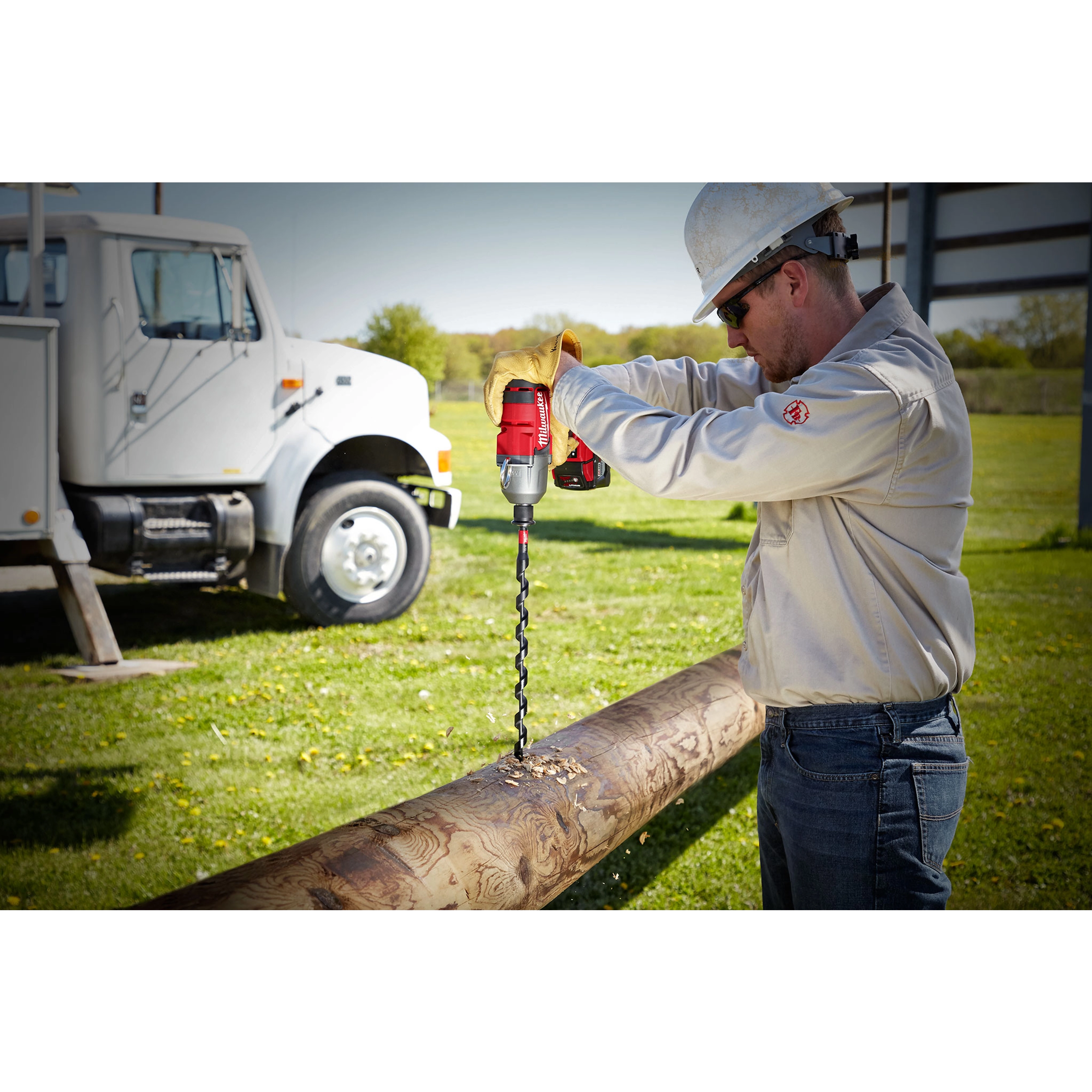 A worker in safety gear uses a red power drill with the SHOCKWAVE™ 13/16" 18" Lineman's Utility Auger Bit to drill into a wooden utility pole. A white utility truck is parked in the background on grassy terrain.