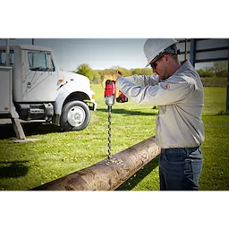 A worker in safety gear uses a red power drill with the SHOCKWAVE™ 13/16" 18" Lineman's Utility Auger Bit to drill into a wooden utility pole. A white utility truck is parked in the background on grassy terrain.