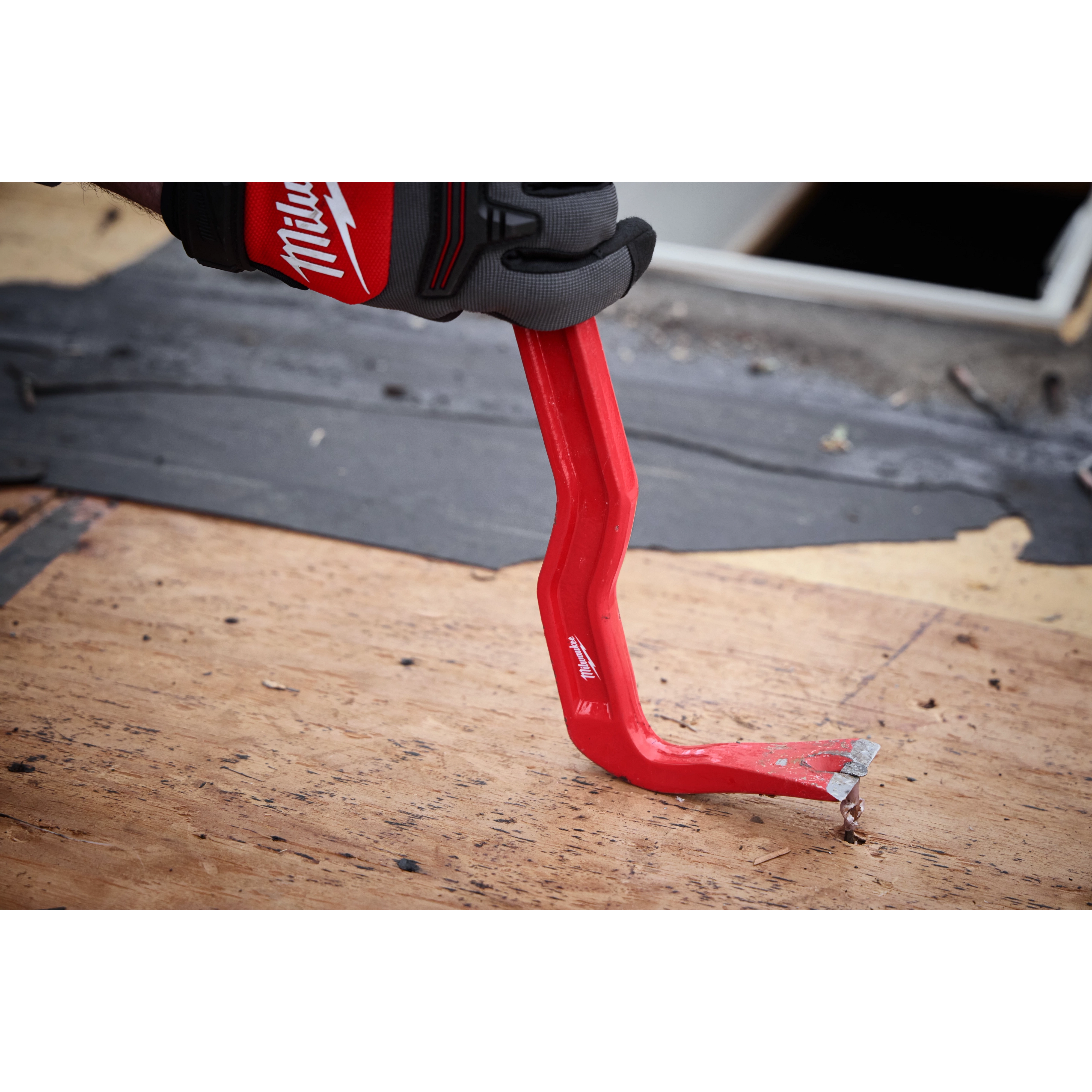 The 18" Wrecking Bar is shown being used to remove nails from a wooden surface. It is bright red with a chiseled end and is held by a hand wearing a Milwaukee-branded glove. The tool is bent for increased leverage and efficiency in demolition tasks.