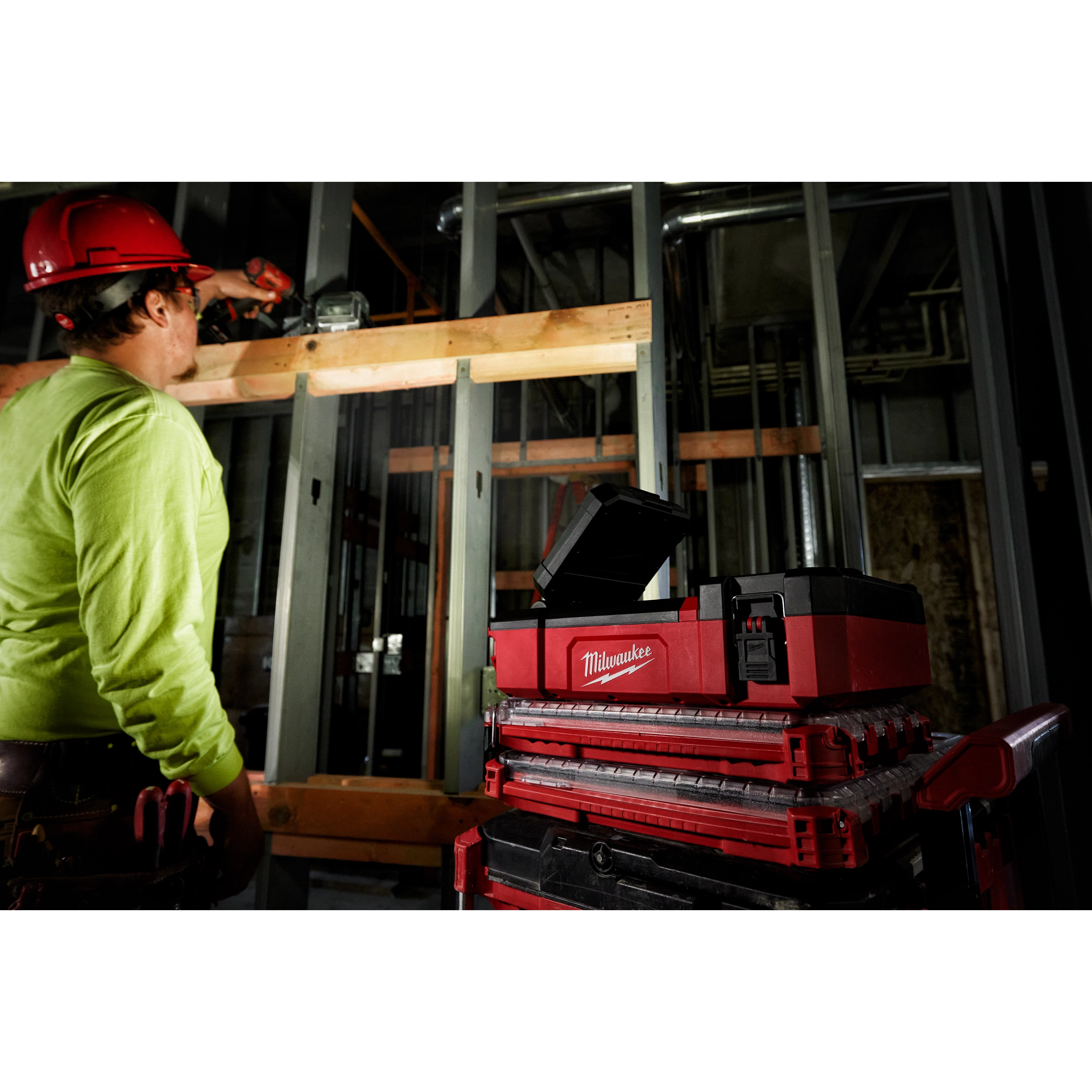 Worker using a drill at a construction site, with M12 PACKOUT Flood Light w/ USB Charging on a stack of red and black toolboxes nearby.