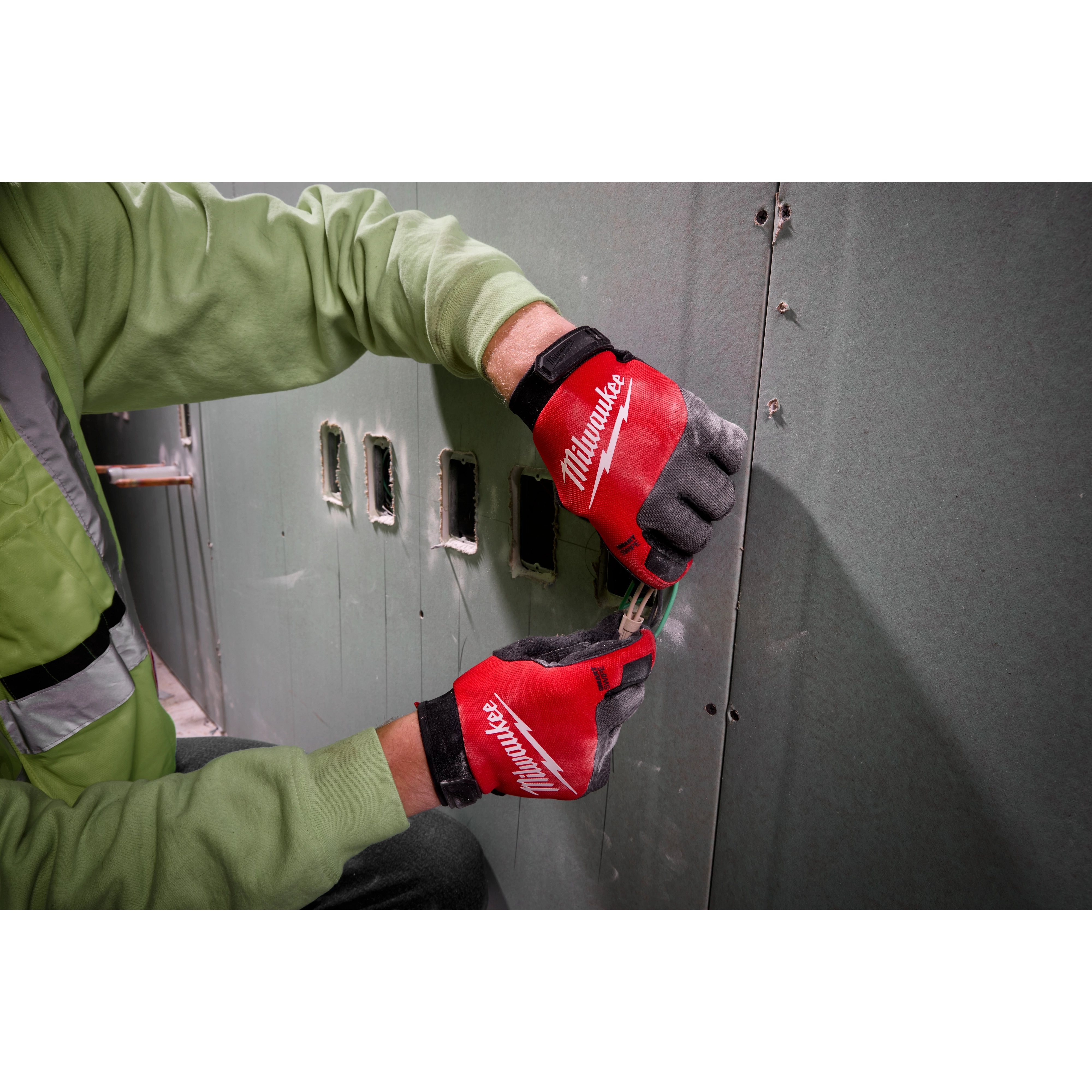 Person wearing Lightweight Work Gloves - L in red and black, working with electrical wires against a green drywall, wearing green shirt.