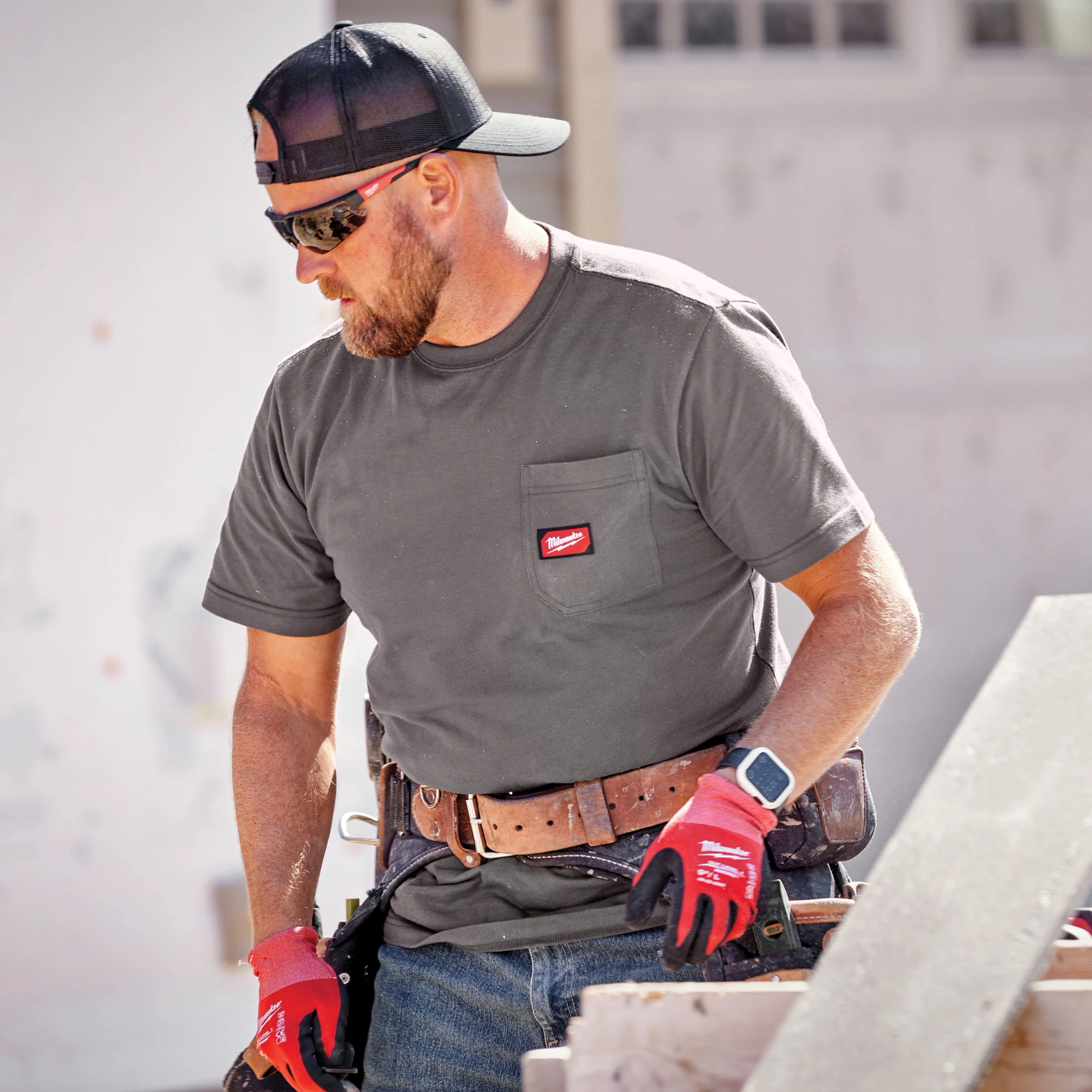Worker wearing a black GRIDIRON Snapback Trucker Hat, grey t-shirt, red gloves, tool belt, and a smartwatch on a construction site.