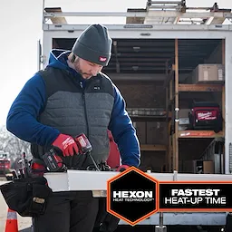 A construction worker wears an M12 Heated AXIS Vest while working with a power tool outside a trailer.