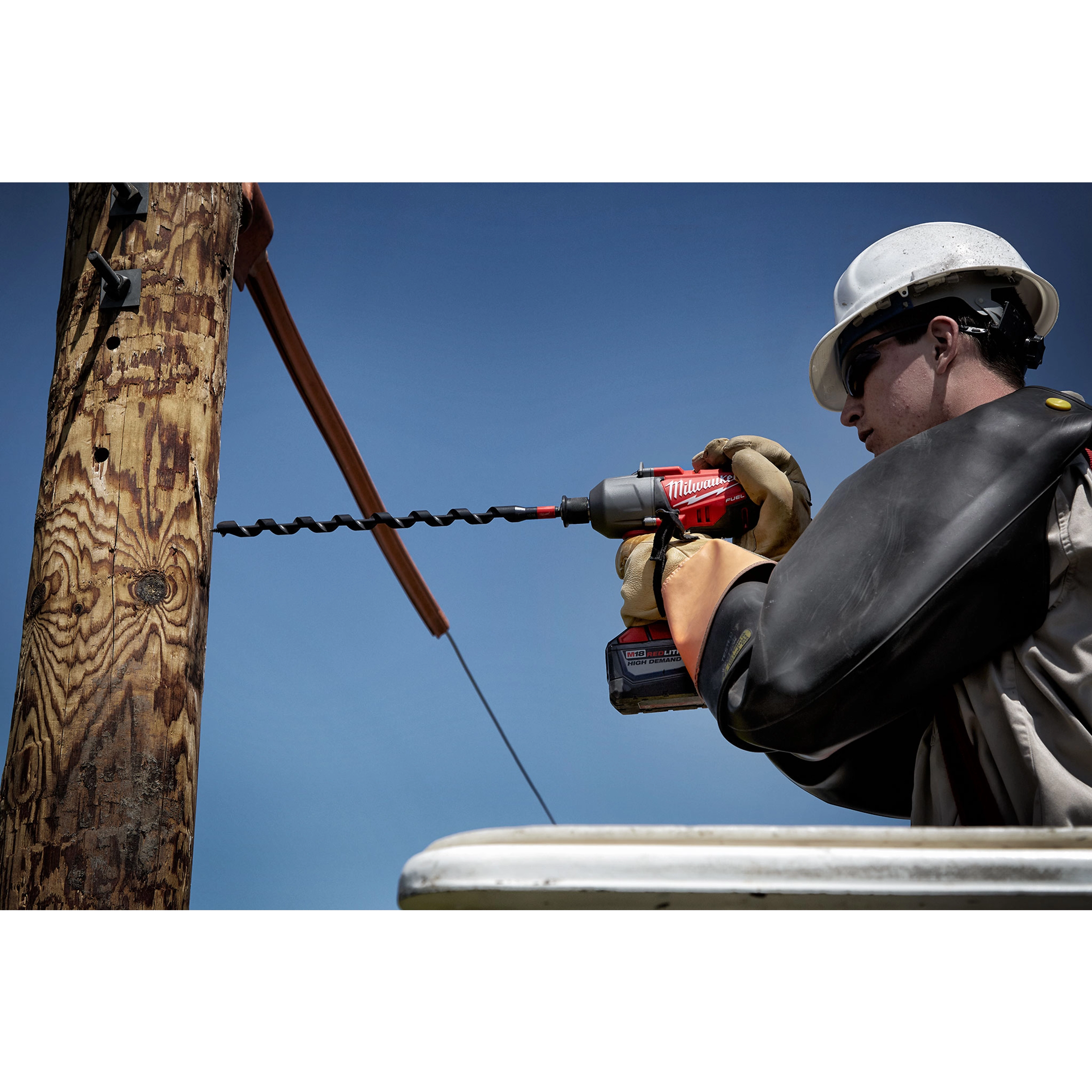 A worker uses the SHOCKWAVE™ 13/16" 18" Lineman's Utility Auger Bit to drill into a wooden utility pole. The bit is attached to a cordless electric drill, and the worker is elevated, wearing protective gloves and a helmet.