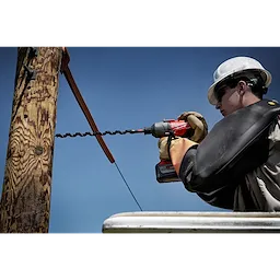A worker uses the SHOCKWAVE™ 13/16" 18" Lineman's Utility Auger Bit to drill into a wooden utility pole. The bit is attached to a cordless electric drill, and the worker is elevated, wearing protective gloves and a helmet.