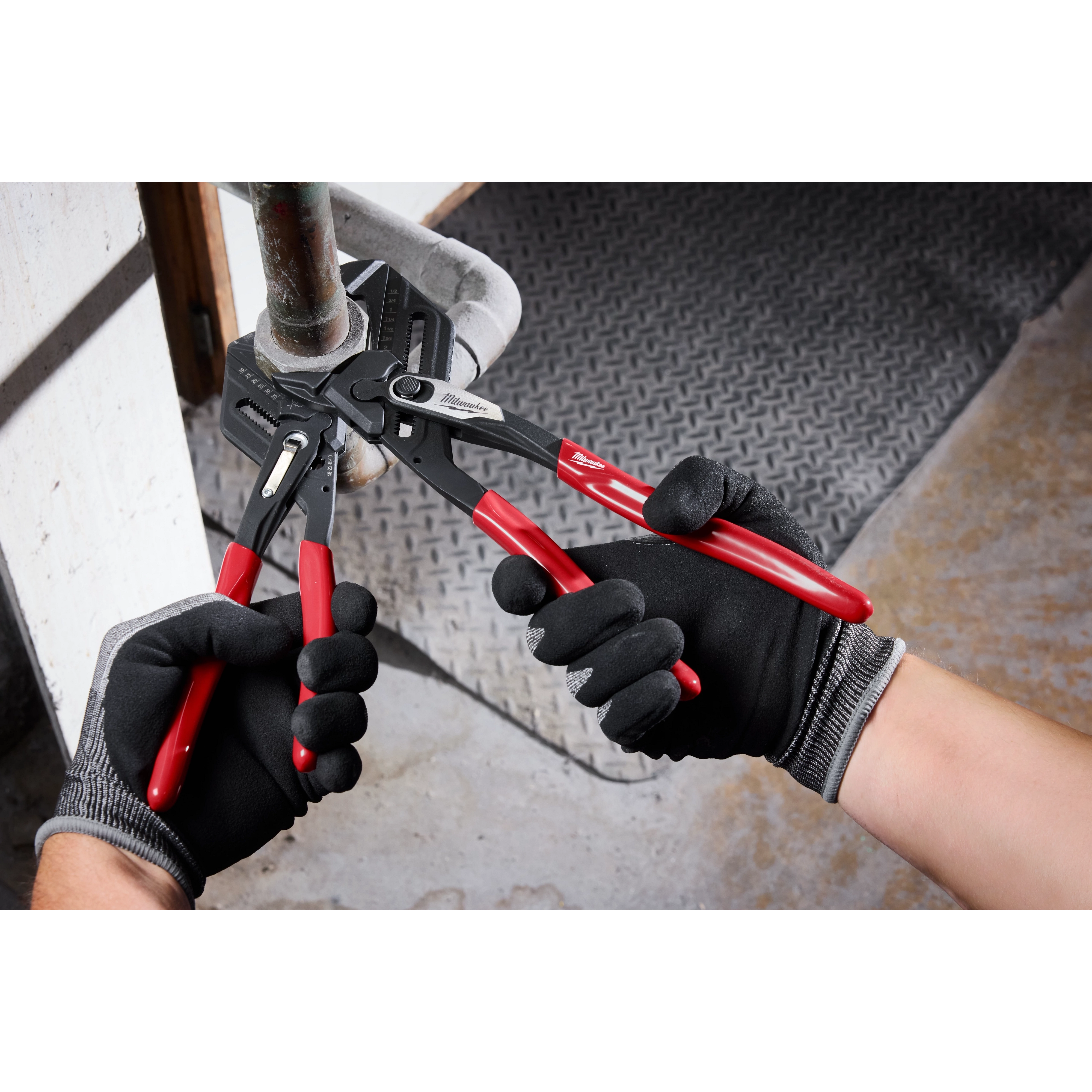Two gloved hands using 10" Pliers Wrench with red and black handles to grip and twist a pipe. The tool is in use in a workshop environment.