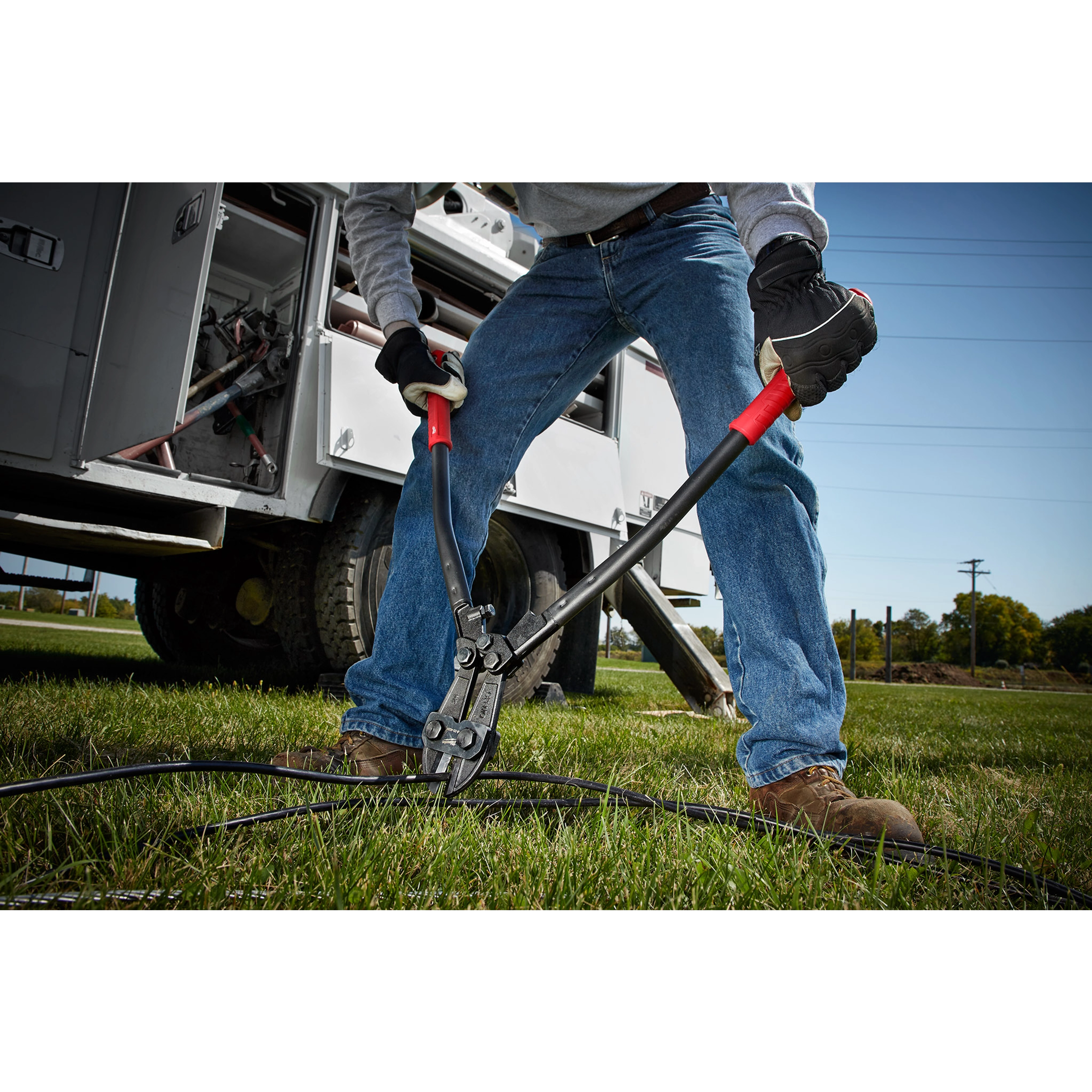 Person using 24" Bolt Cutter with red handles to cut a thick black cable on grassy ground, near a work truck.
