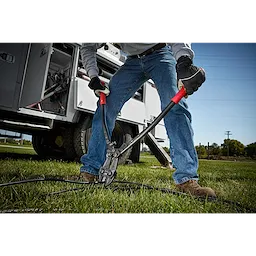 Person using 24" Bolt Cutter with red handles to cut a thick black cable on grassy ground, near a work truck.